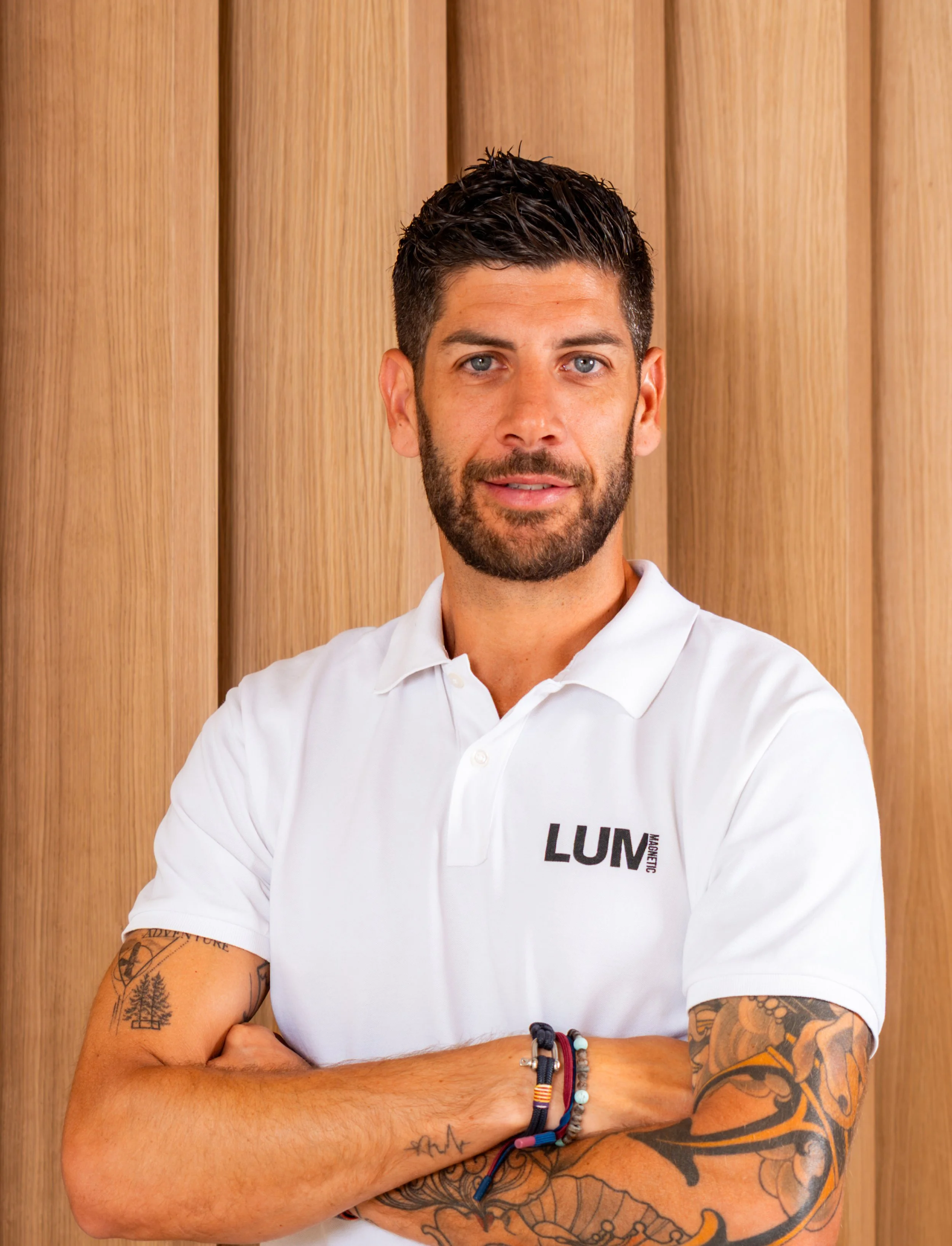 Un hombre con barba y cabello oscuro, con tatuajes en ambos brazos, lleva puesta una camiseta blanca con el logo 'LUM' y está de brazos cruzados frente a una pared de madera.