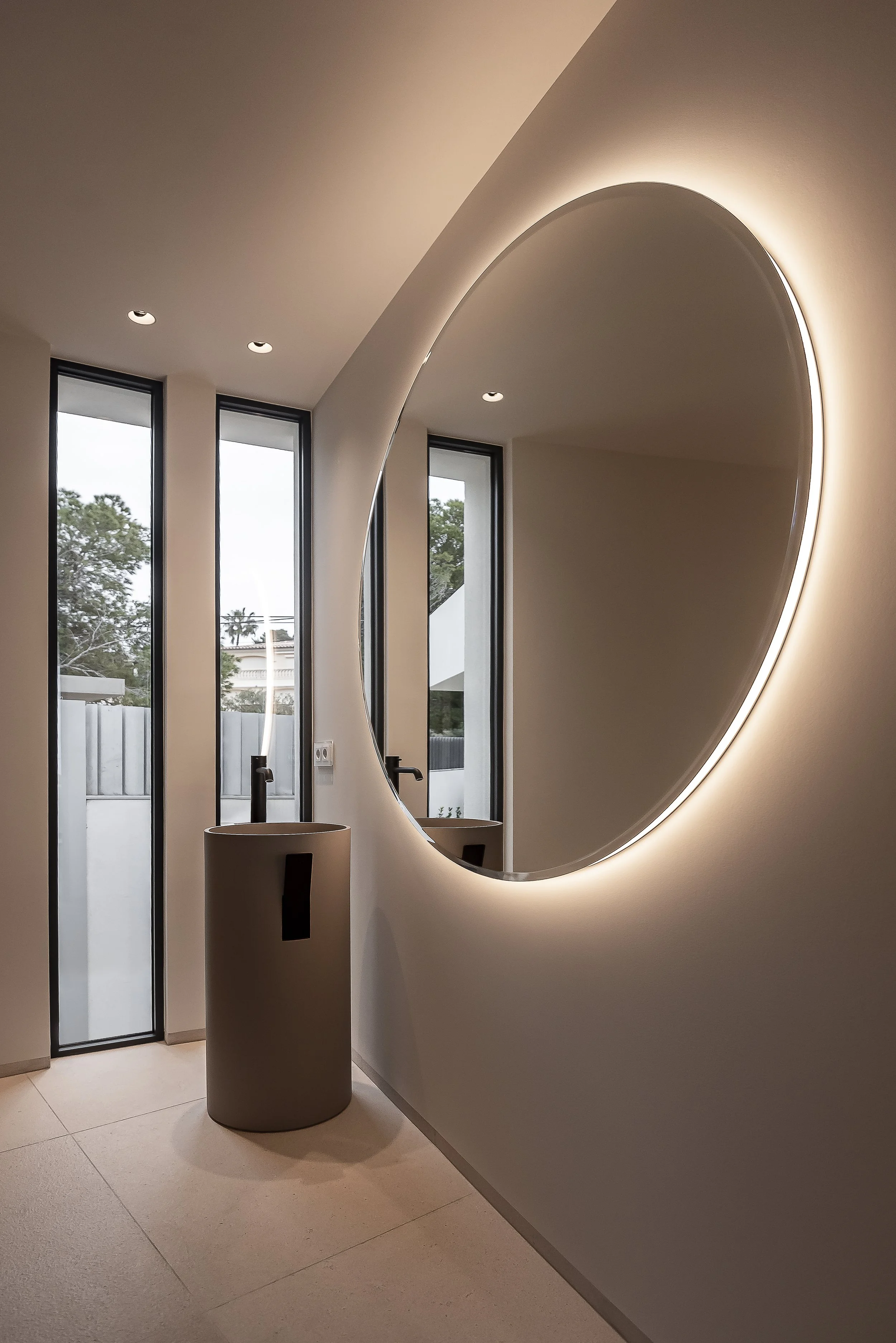 Espejo redondo con iluminación de fondo en una pared de interior, junto a ventanas altas y una lámpara en un espacio moderno.