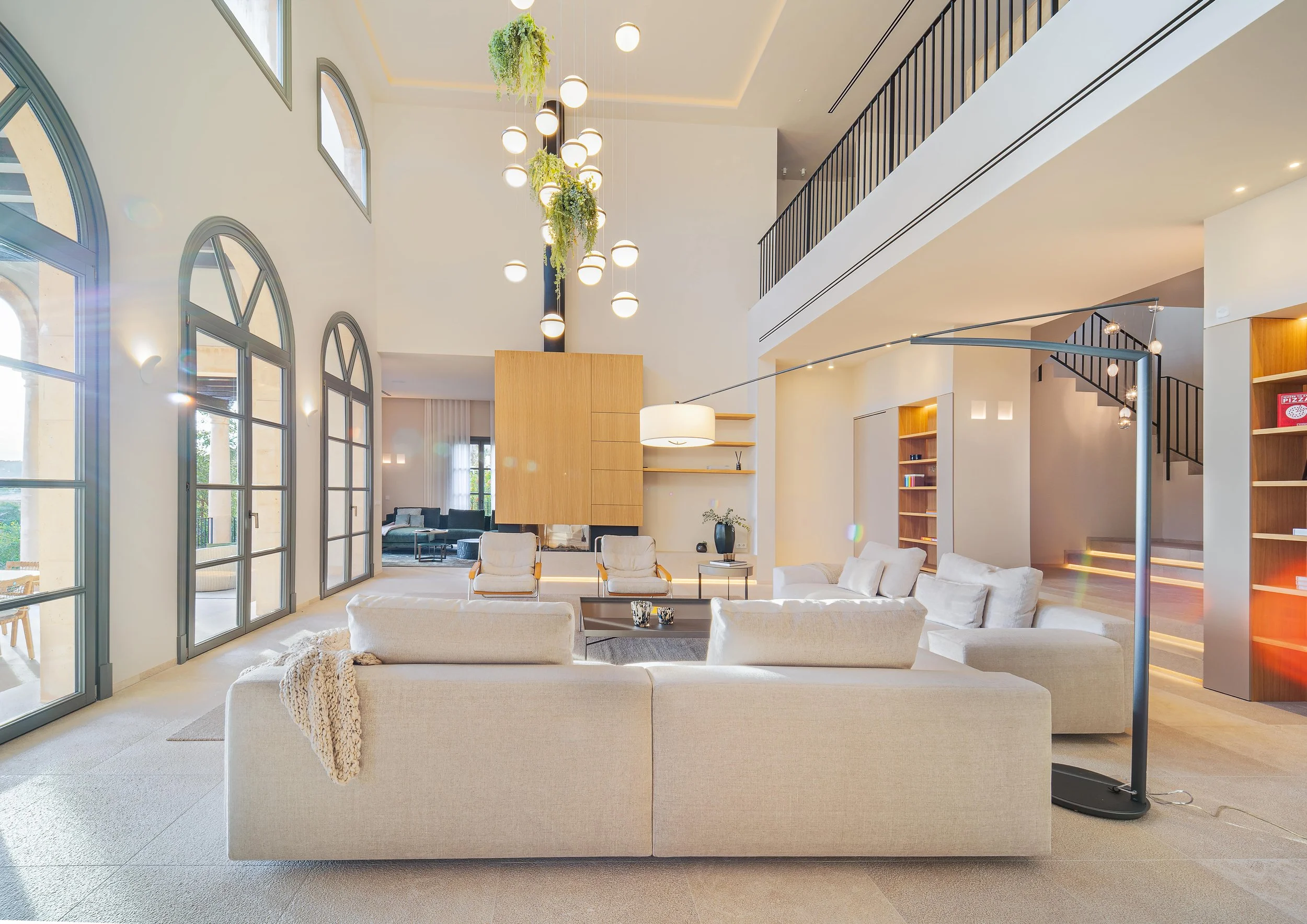Interior moderno de sala con sillones blancos, mesa de café negra, libreros de madera y escalera de herrería negra. Grandes ventanas de arcos y lámparas colgantes con plantas verdes.
