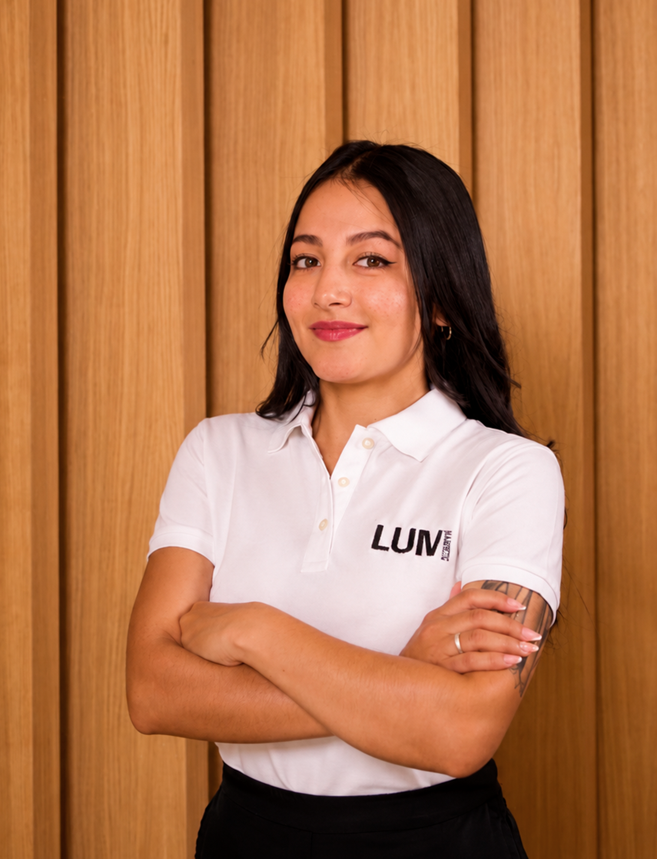 Joven mujer con cabello oscuro, vestida con camiseta blanca con logo de LUM, de brazos cruzados, posando frente a fondo de madera.