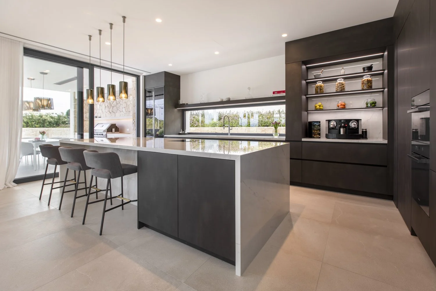 Cocina moderna con isla de granito, gabinetes oscuros, barra con cinco sillas y una área de coffee en el estante abierto con máquinas de café, frutas y alimentos en frascos de vidrio.