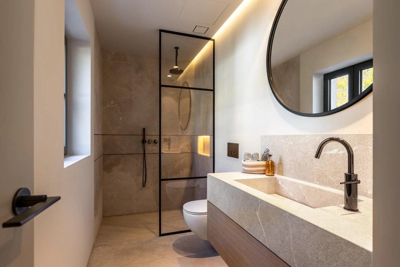 Baño moderno con lavabo de piedra, espejo grande redondo, inodoro, ducha con mampara de vidrio y grifería negra, ventana con vista a árboles.