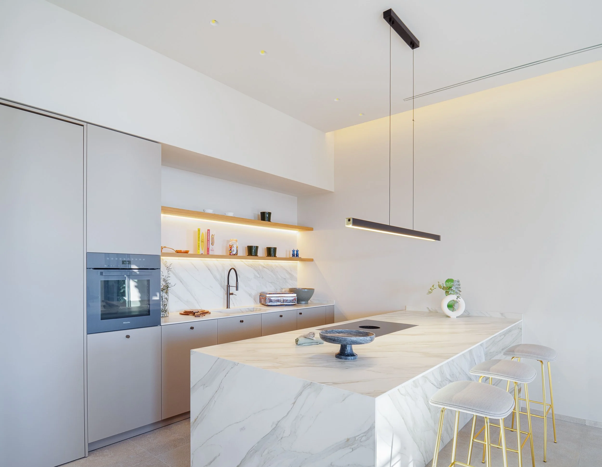 Cocina moderna con armarios blancos, encimera de mármol, estantería con decoración y barandales con sillas. Colores neutros y diseño minimalista.