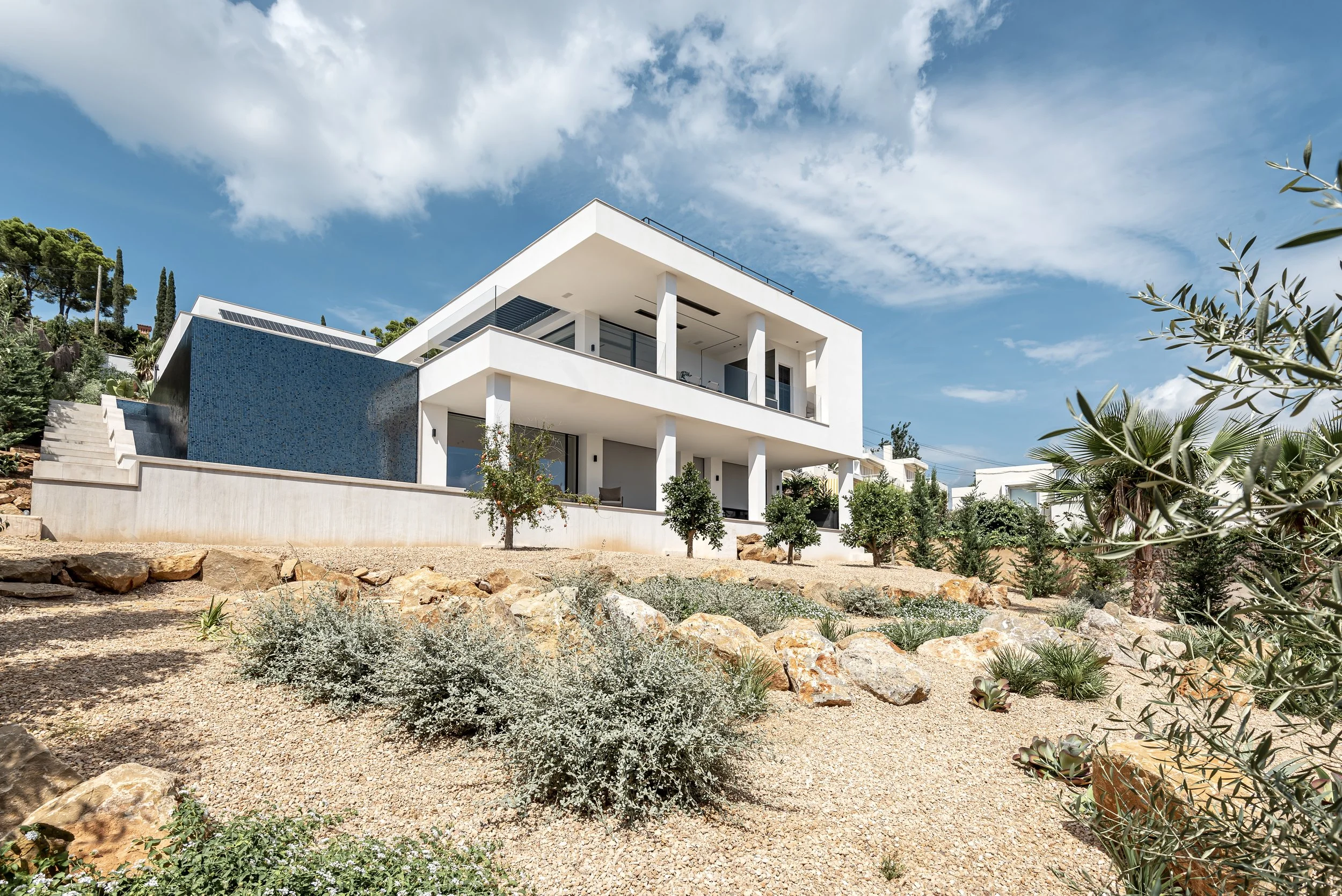 Casa moderna de dos pisos con balcones y piscina en un entorno soleado con plantas y rocas en primer plano, cielo nublado parcialmente.