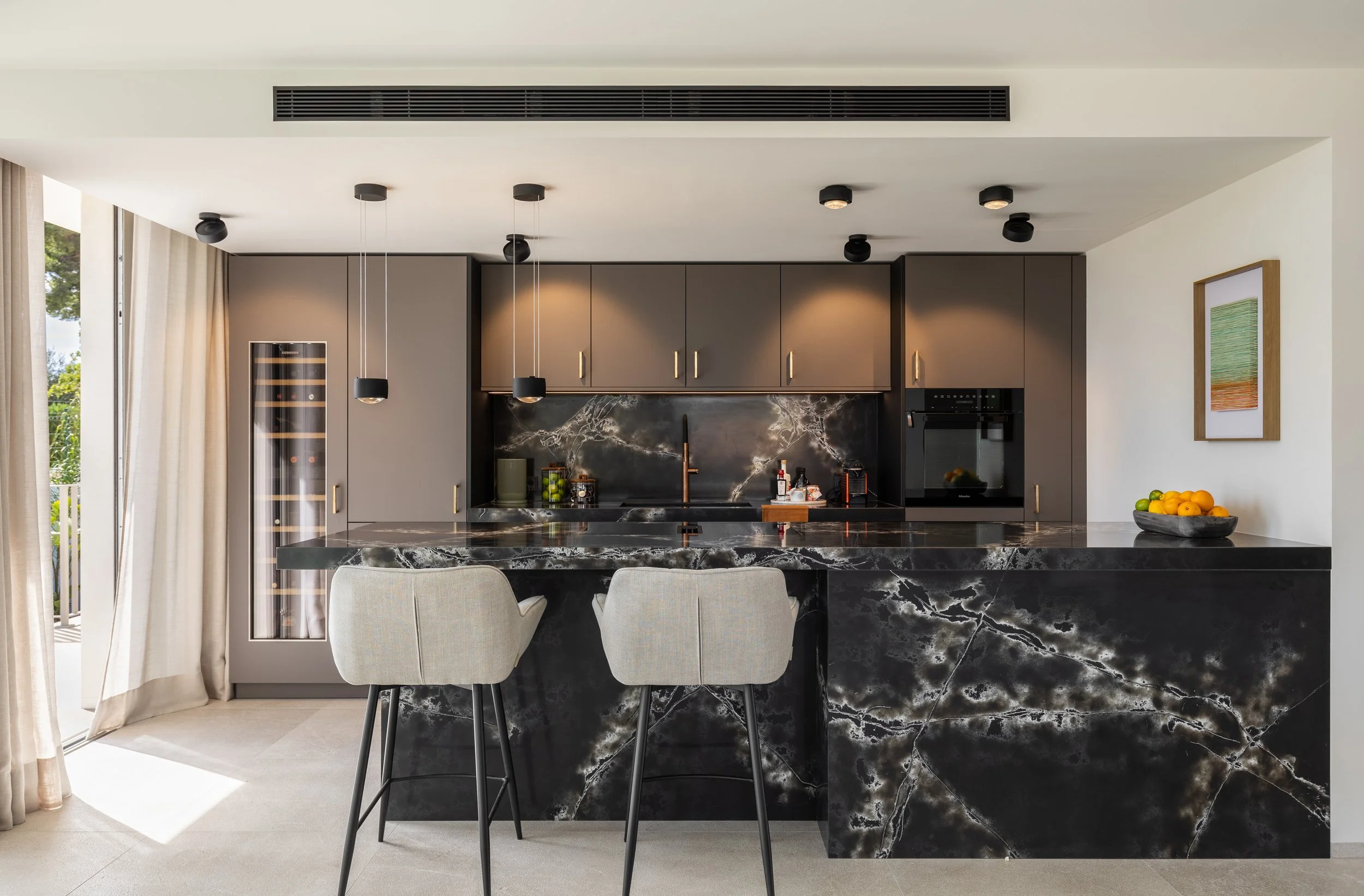 Cocina moderna con gabinetes grises, encimera y espalda de mármol oscuro, lámparas negras en el techo, ventanas con cortinas blancas a la izquierda, frutas en un tazón y un cuadro con líneas de colores en la pared derecha.