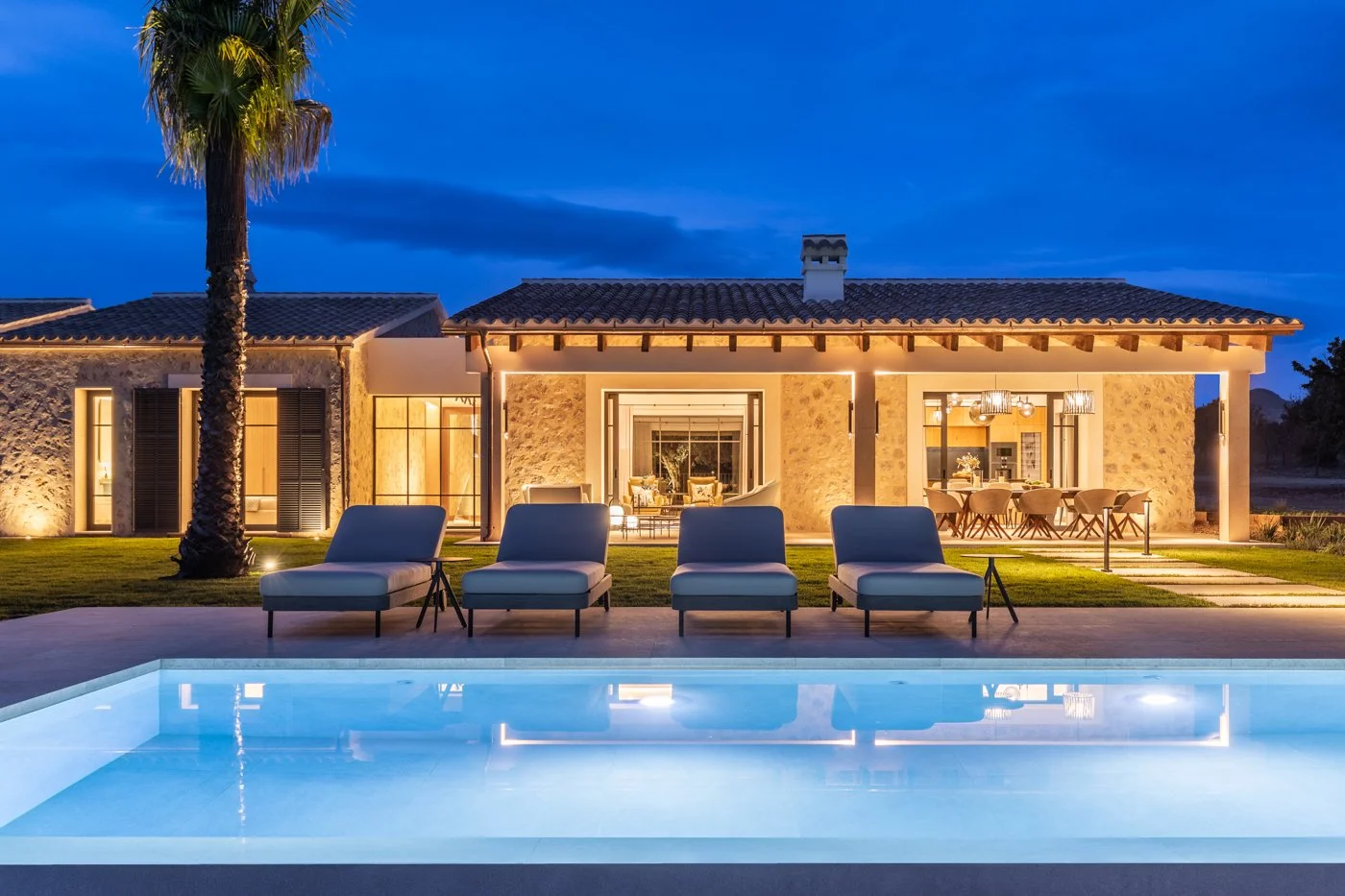 Casa moderna con iluminación cálida, piscina y tumbonas en primer plano, en un entorno nocturno.
