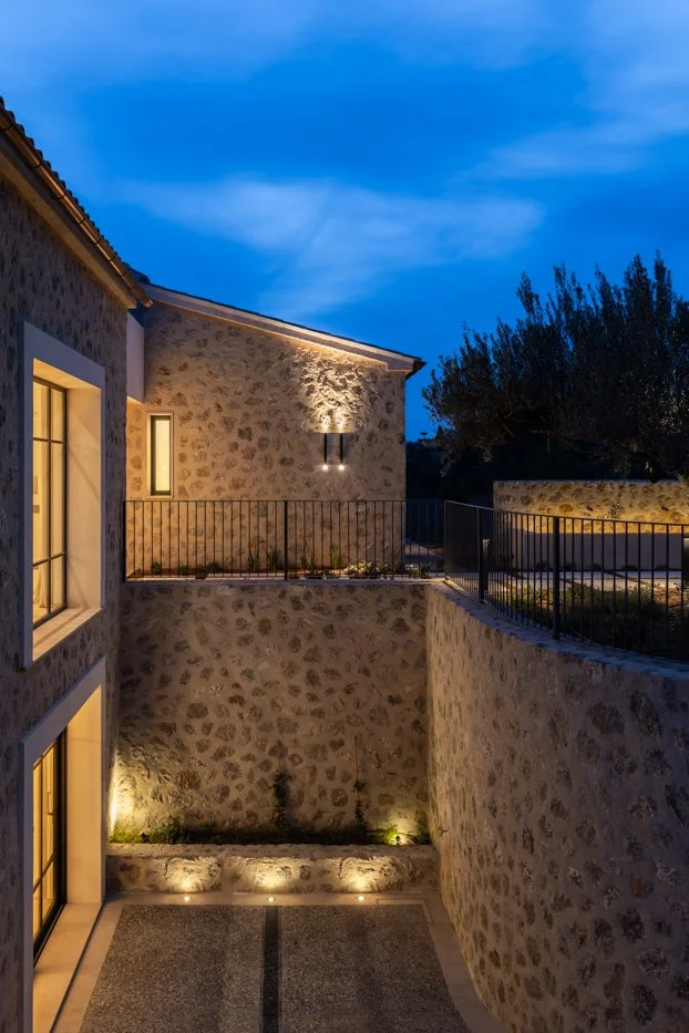 Casa de piedra con luces exteriores en la noche, vista desde un patio con paredes de piedra y barandales negros.