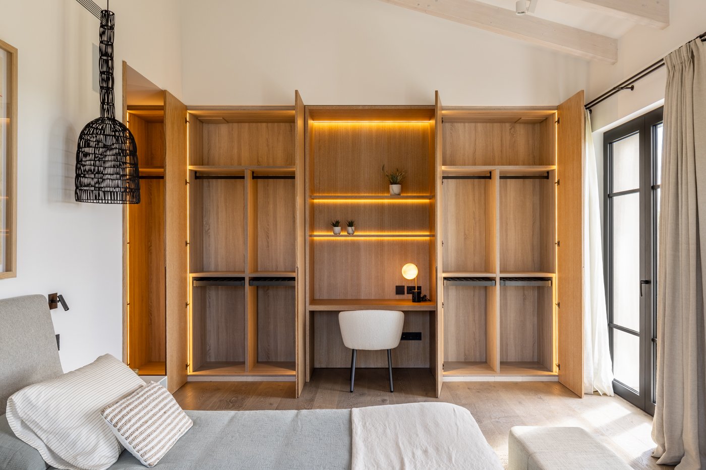 Habitación moderna con clóset de madera y luz led, escritorio con lámpara, sillón, cama con almohadas, cortinas y ventana con puerta de vidrio.