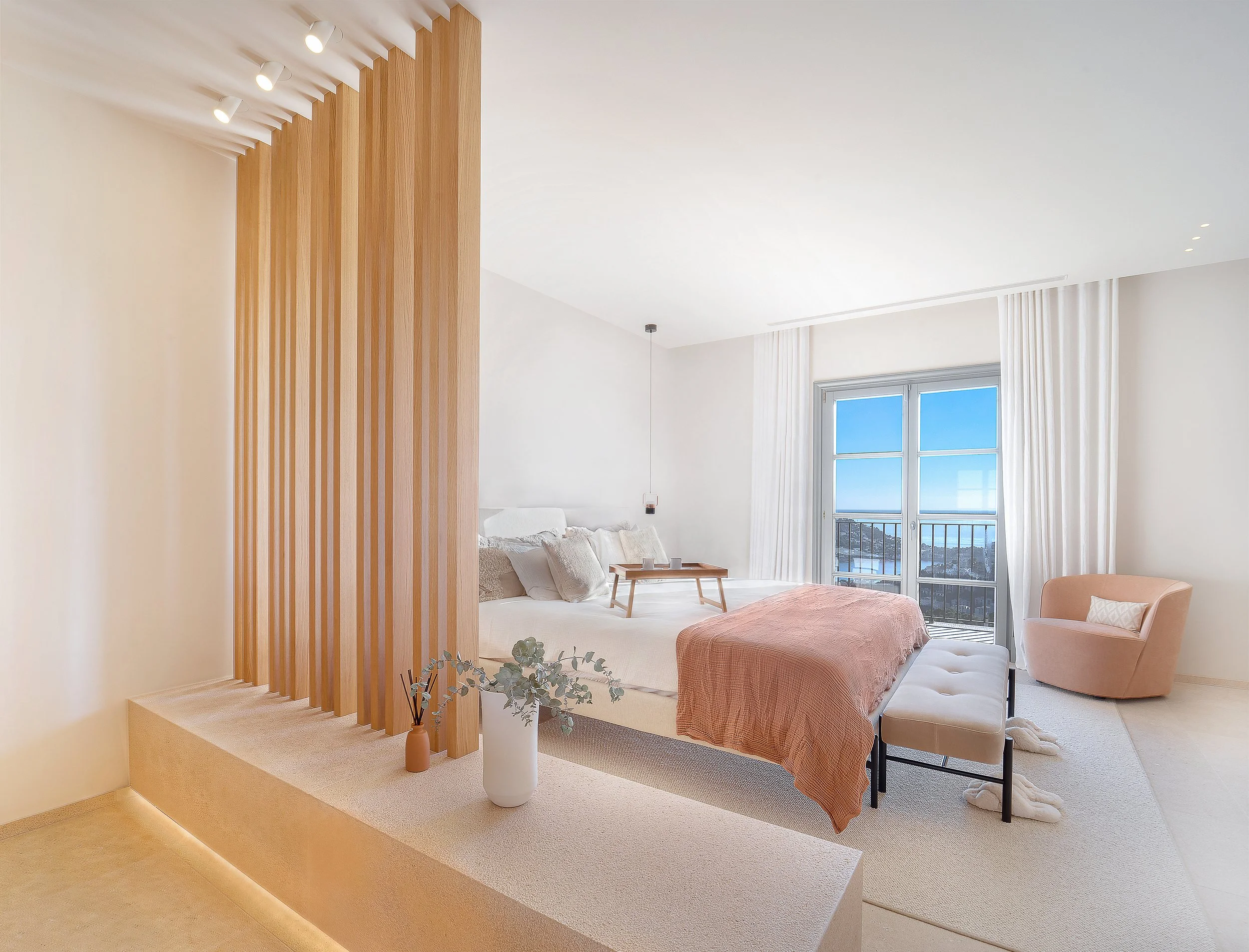 Sala de estar luminosa con muebles en tonos neutros y accesorios decorativos, vista hacia una ventana grande con cortinas blancas y una vista del mar en el fondo.