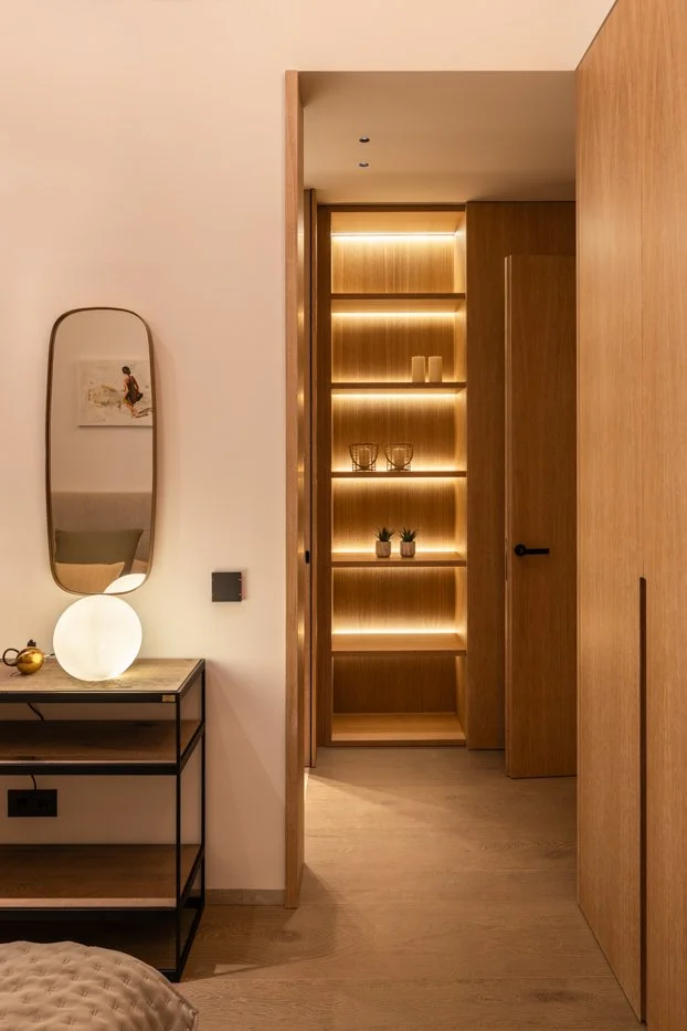 Vista de un espacio interior con estantería de madera iluminada, un espejo de forma irregular, una mesa con lámpara de mesa y una pared de tonos claros.
