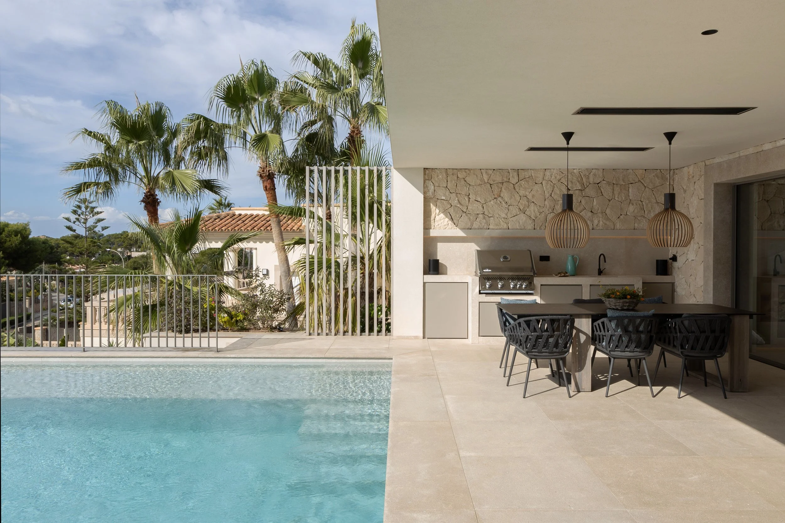 Piscina en un patio con mesas y sillas, palmeras y una casa moderna con paredes de piedra y techo blanco bajo un cielo despejado.
