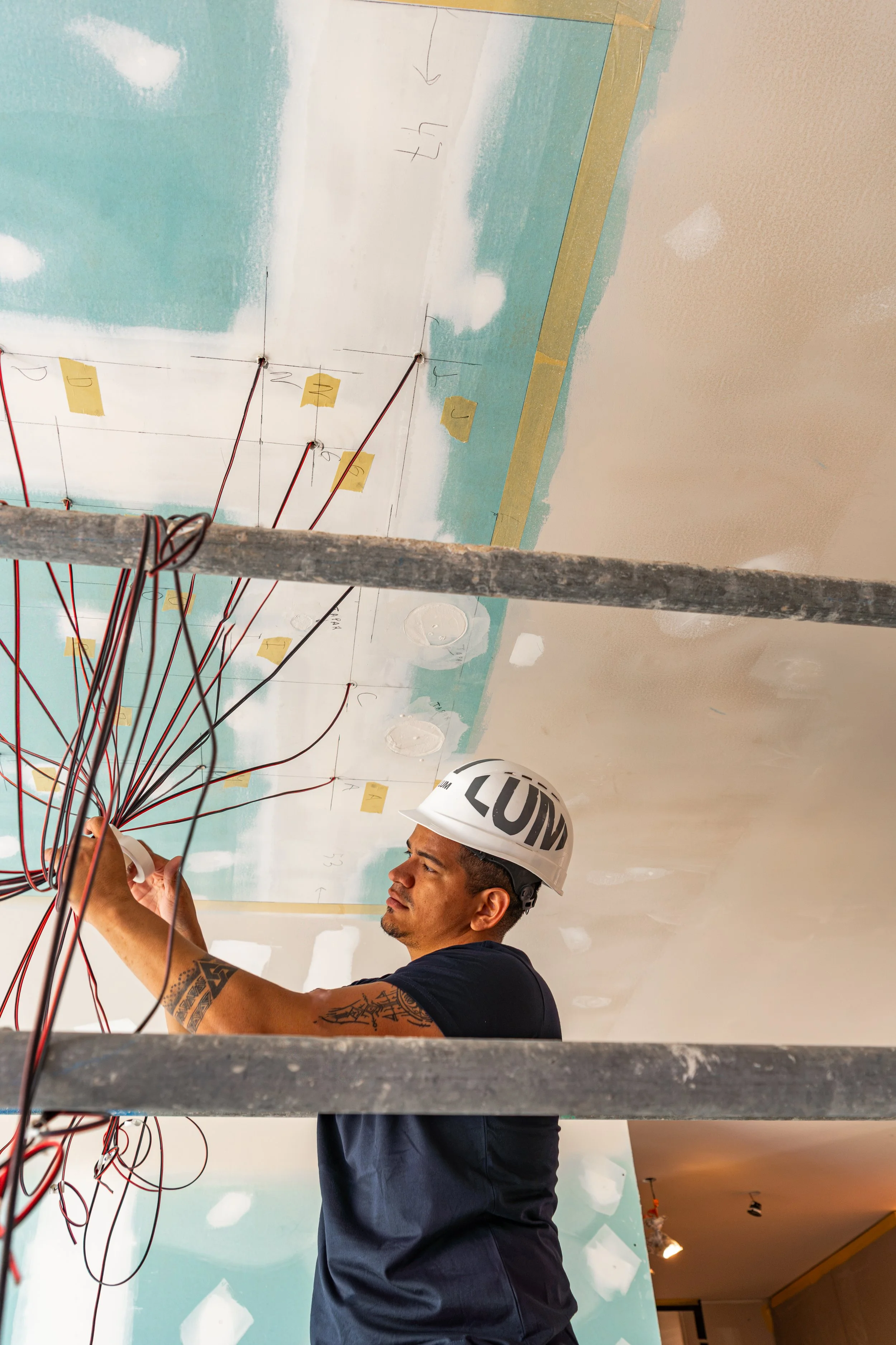 Un hombre con casco blanco que tiene las letras en inglés 'LUM' y tatuajes en un brazo, trabaja en la instalación de cables eléctricos en un techo en construcción.