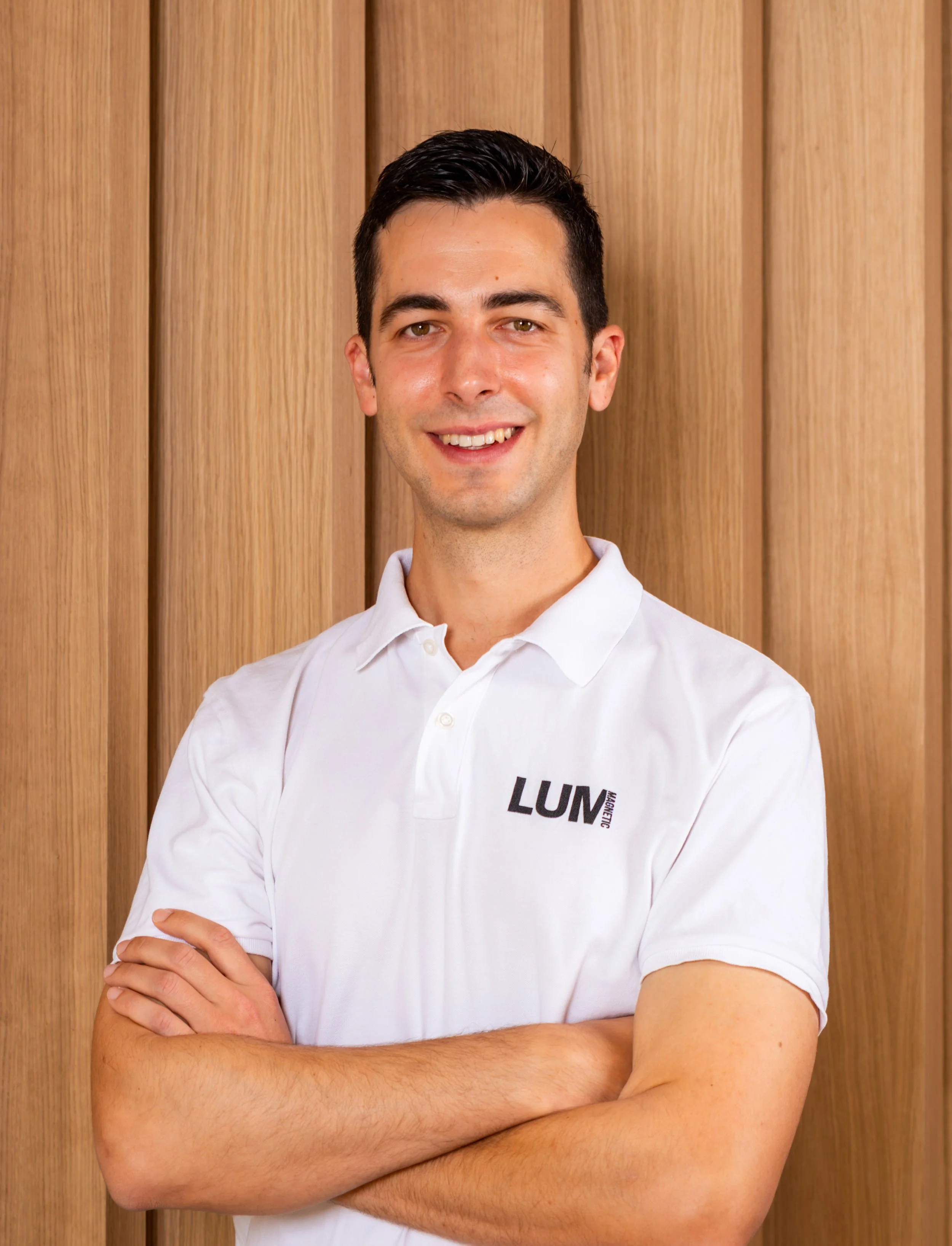 Joven hombre con camisa blanca con letras negras que dicen 'LUM' en la parte izquierda del pecho, con los brazos cruzados, posando frente a una pared de madera clara.