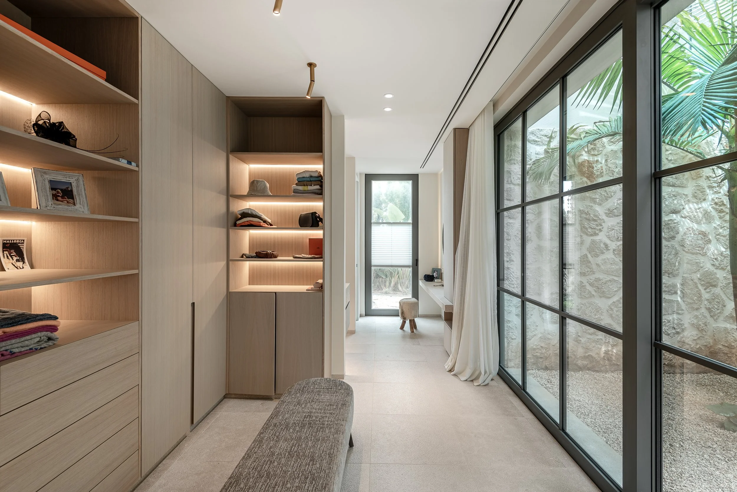 Interior de una sala con paredes de madera y piso de baldosas, estantes con ropa doblada, ventana grande con cortinas blancas, puerta de vidrio y paredes de piedra decorativa, con un asiento de tela y vista de plantas exteriores.