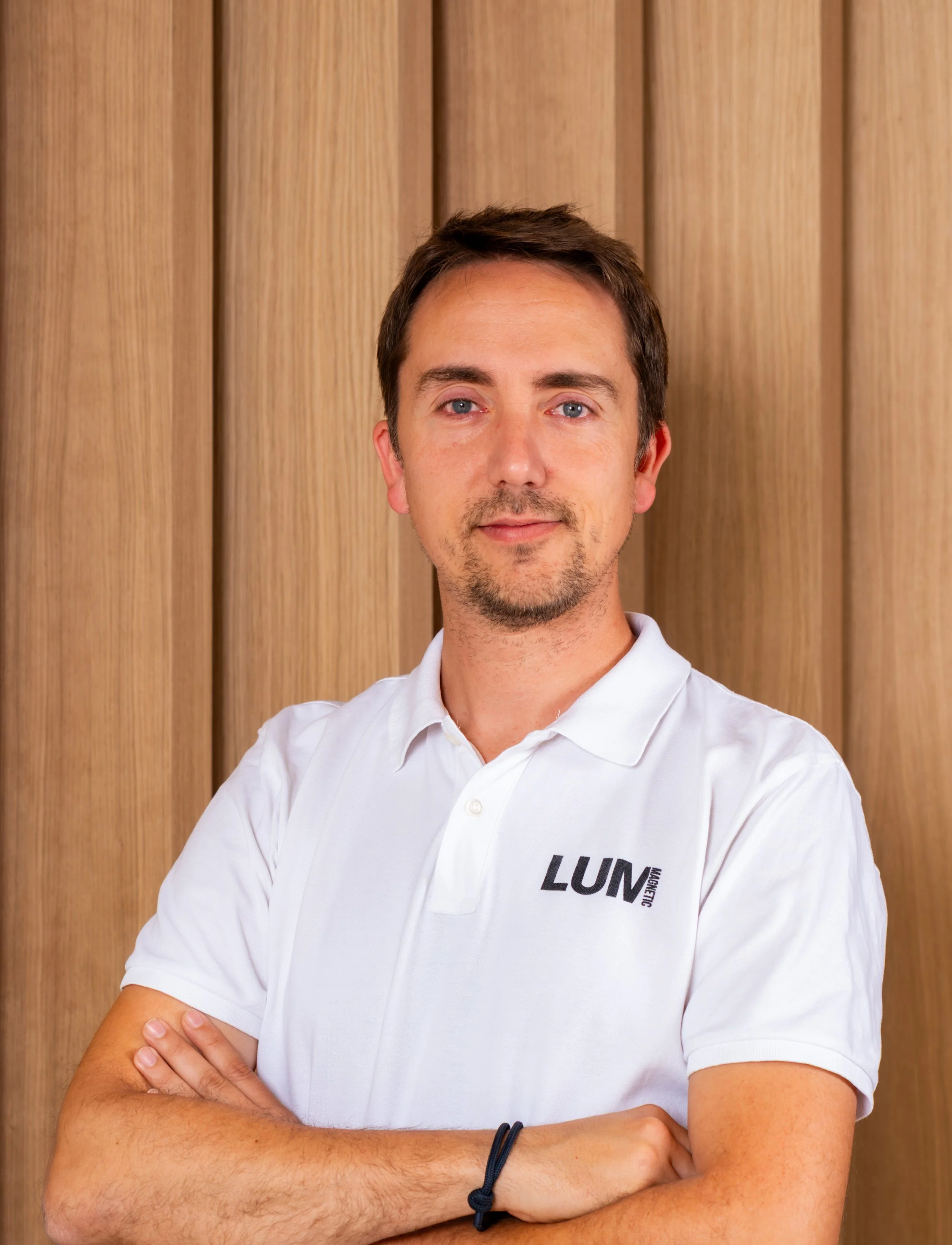 Primer plano de un hombre con barba y cabello castaño, con camiseta blanca con logotipo de LUM, de brazos cruzados, frente a fondo de madera.