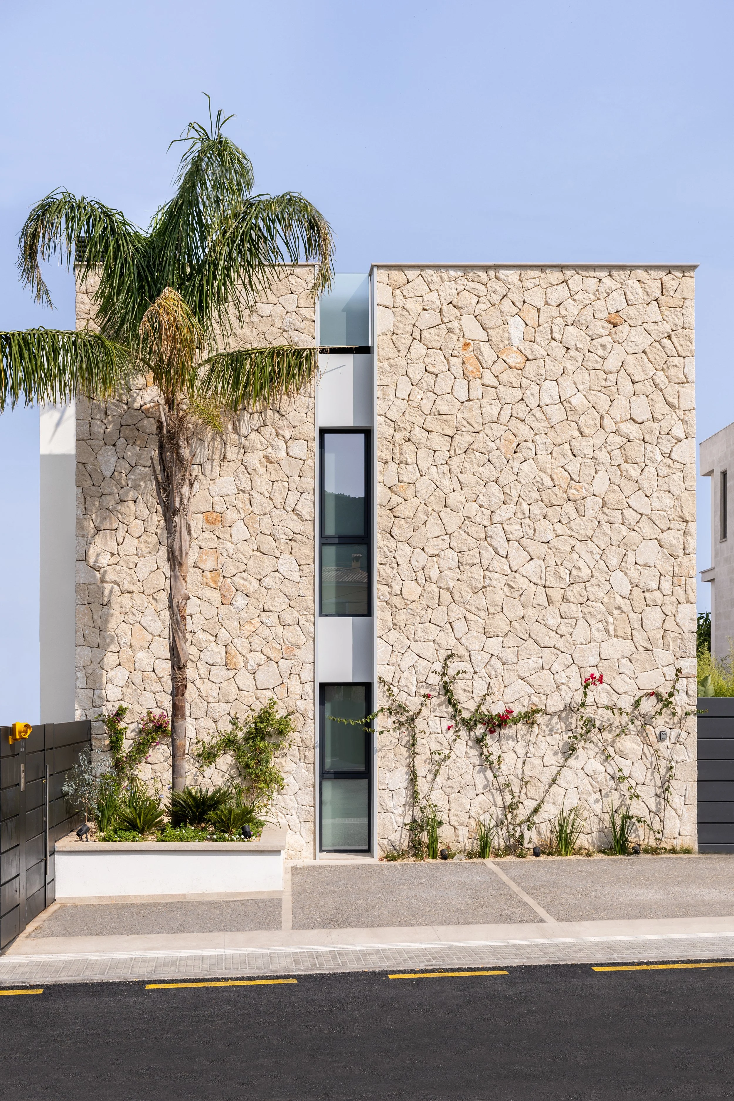 Fachada de un edificio moderno con paredes de piedra, ventanas altas y delgadas, y un árbol de palma frente al edificio. En primer plano, la calle con acera y líneas de tránsito.