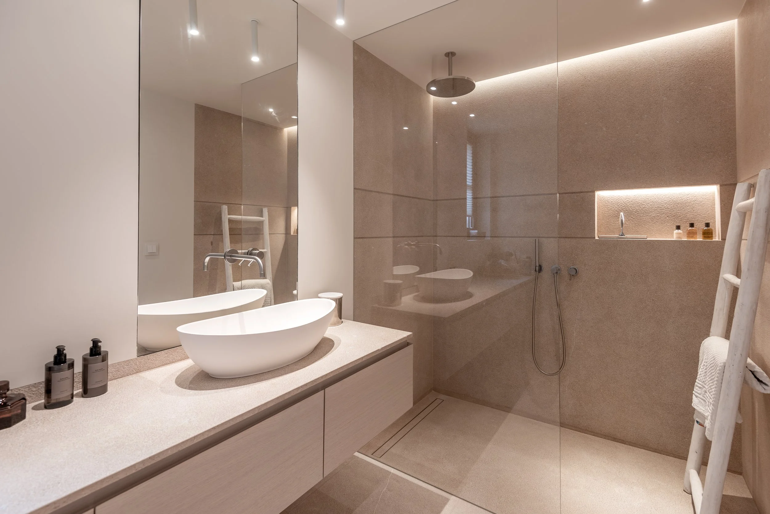 Baño moderno con espejo grande, lavabo ovalado en encimera de piedra, botellas de jabón, ducha con paredes de piedra, accesorios blancos y una escalera de madera