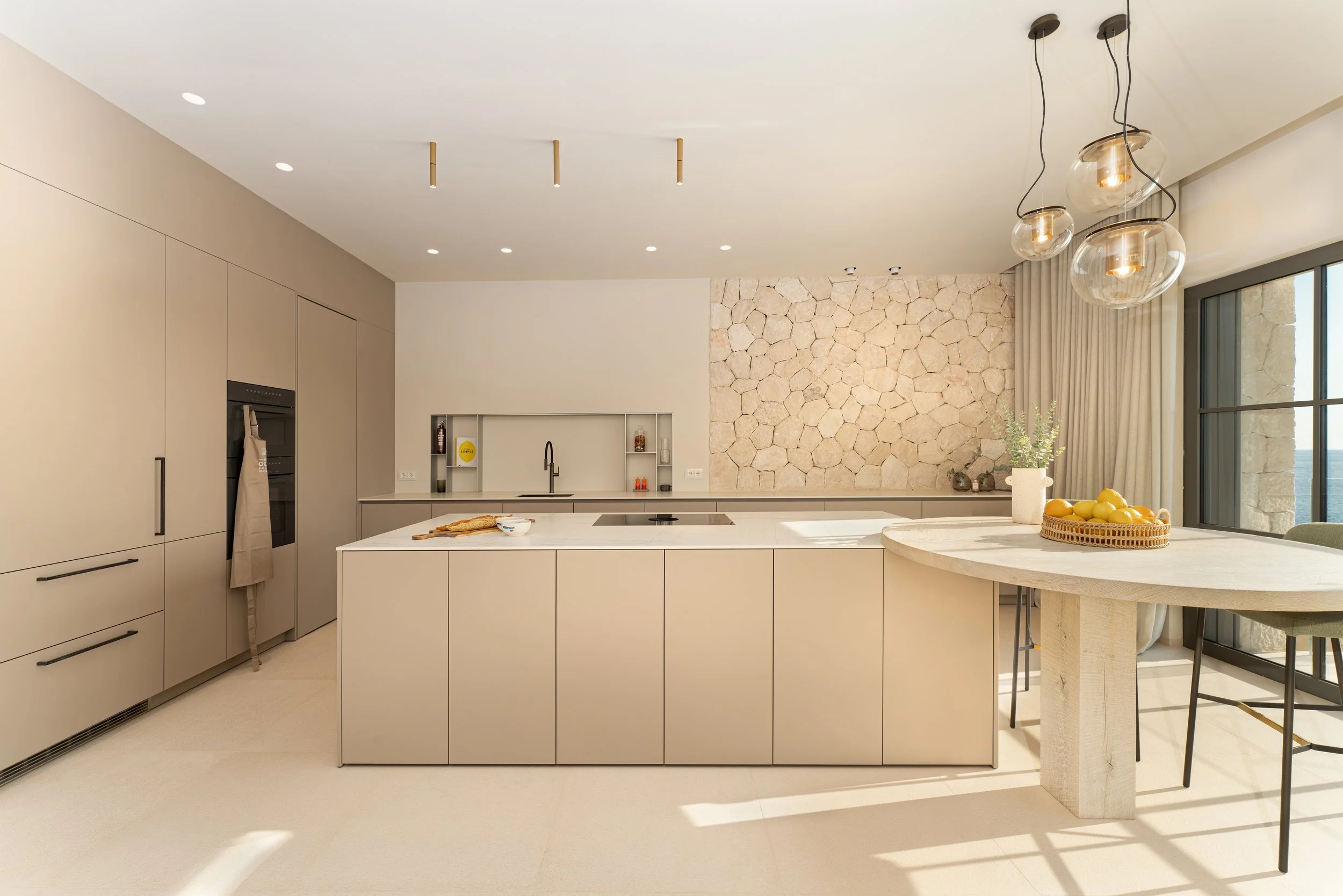 Cocina moderna con paredes de piedra, gabinetes beige y una isla central con estufa, decorada con frutas y utensilios de cocina, con una ventana grande y cortinas.