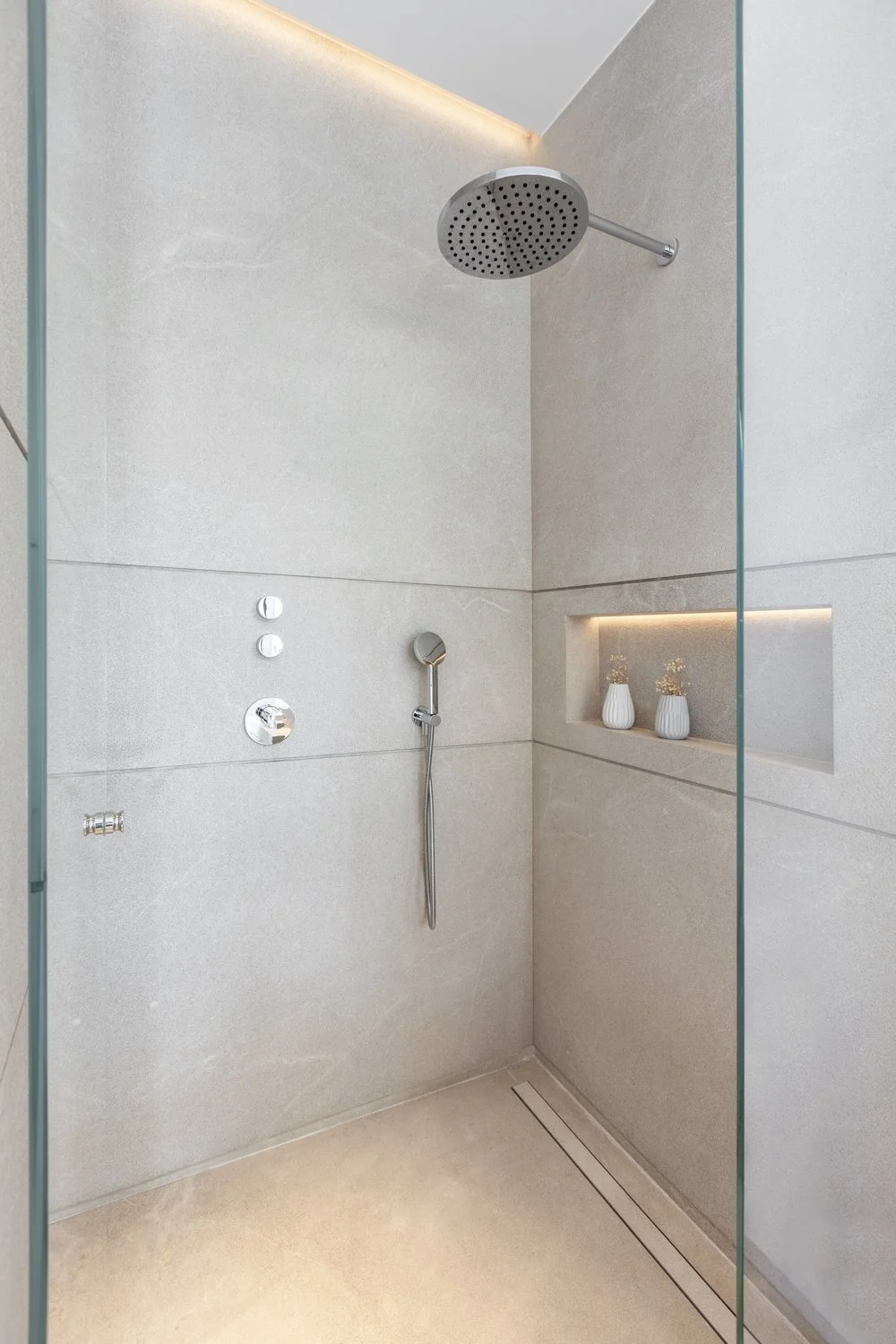 Ducha moderna con cabezal de lluvia, paredes de azulejo gris claro, nicho empotrado con dos jarrones decorativos y luz LED, control de agua y accesorio de mano.