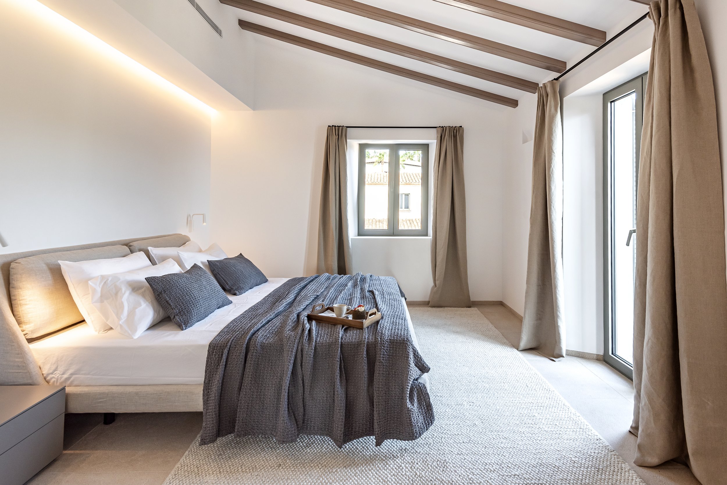 Habitación con cama doble, almohadas blancas y grises, cortinas beige, ventana con vista a los techos de las casas cercanas, paredes blancas, techos con vigas de madera, alfombra Beige y muros limpios y ordenados.