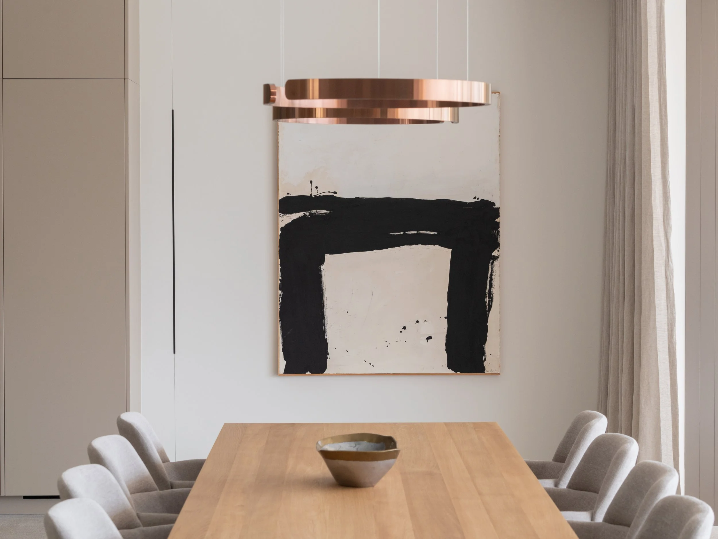 Sala de comedor con mesa de madera y sillas tapizadas, pintura moderna en la pared, cortinas claras en la ventana, cuadro abstracto en la pared y lámpara de diseño en el techo.