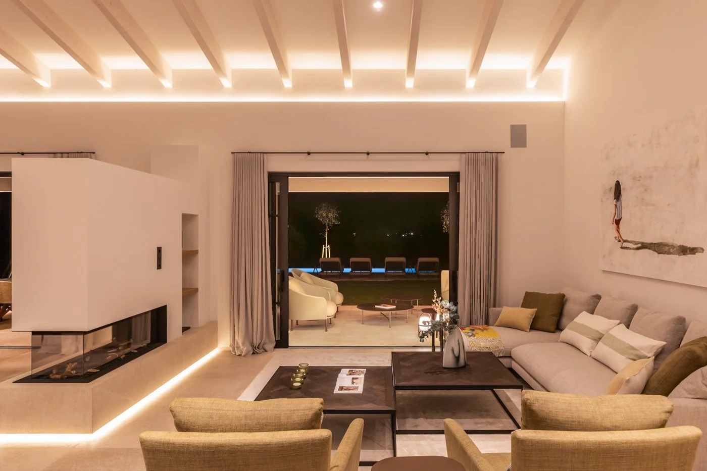 Sala de estar moderna con sofá gris, sillones beige, mesitas de centro, chimenea decorativa, cortinas beige, y balcón con sillas y mesas exteriores, iluminada con luz ambiental cálida.