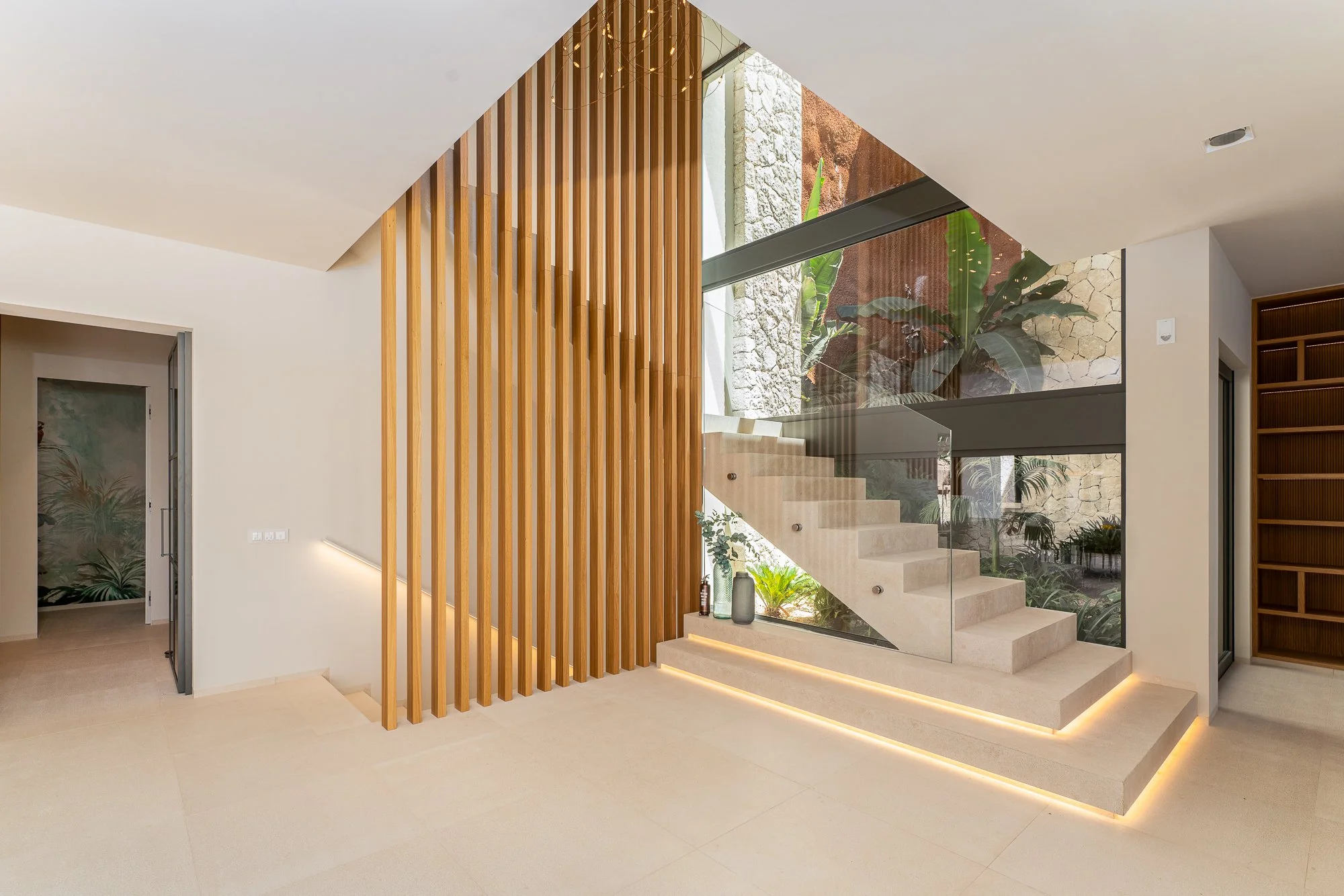Interior modern y brillante de una casa con escalera de piedra y barandillas de vidrio, decorada con plantas y madera tallada, con un muro de piedra y una ventana grande que permite la entrada de luz natural.