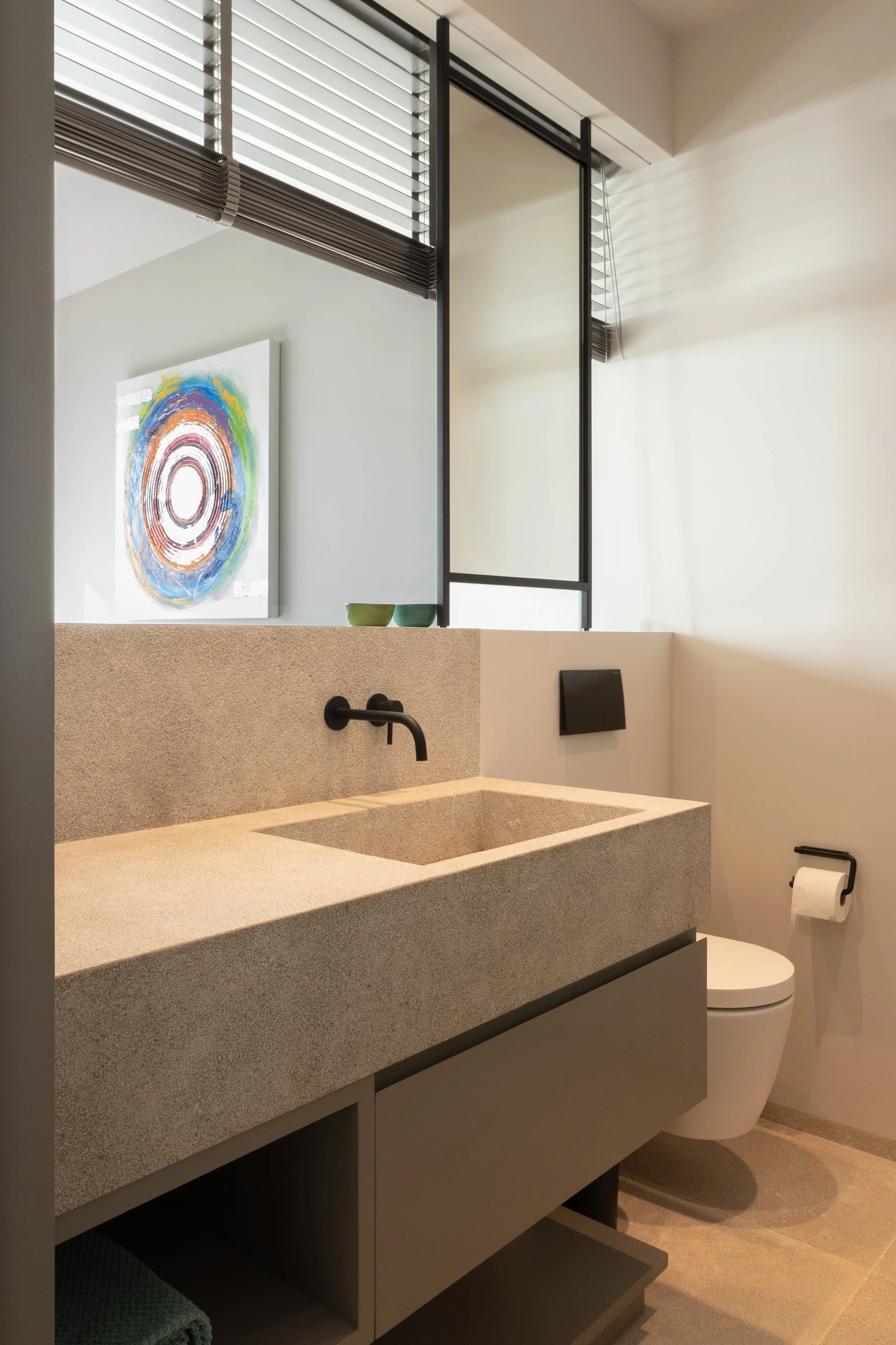 Vista de un baño moderno con lavabo de piedra, grifo negro, arte abstracto colorido en la pared, ventanas con persianas de madera, inodoro blanco, porta papel higiénico, y accesorios en tonos gris y negro.