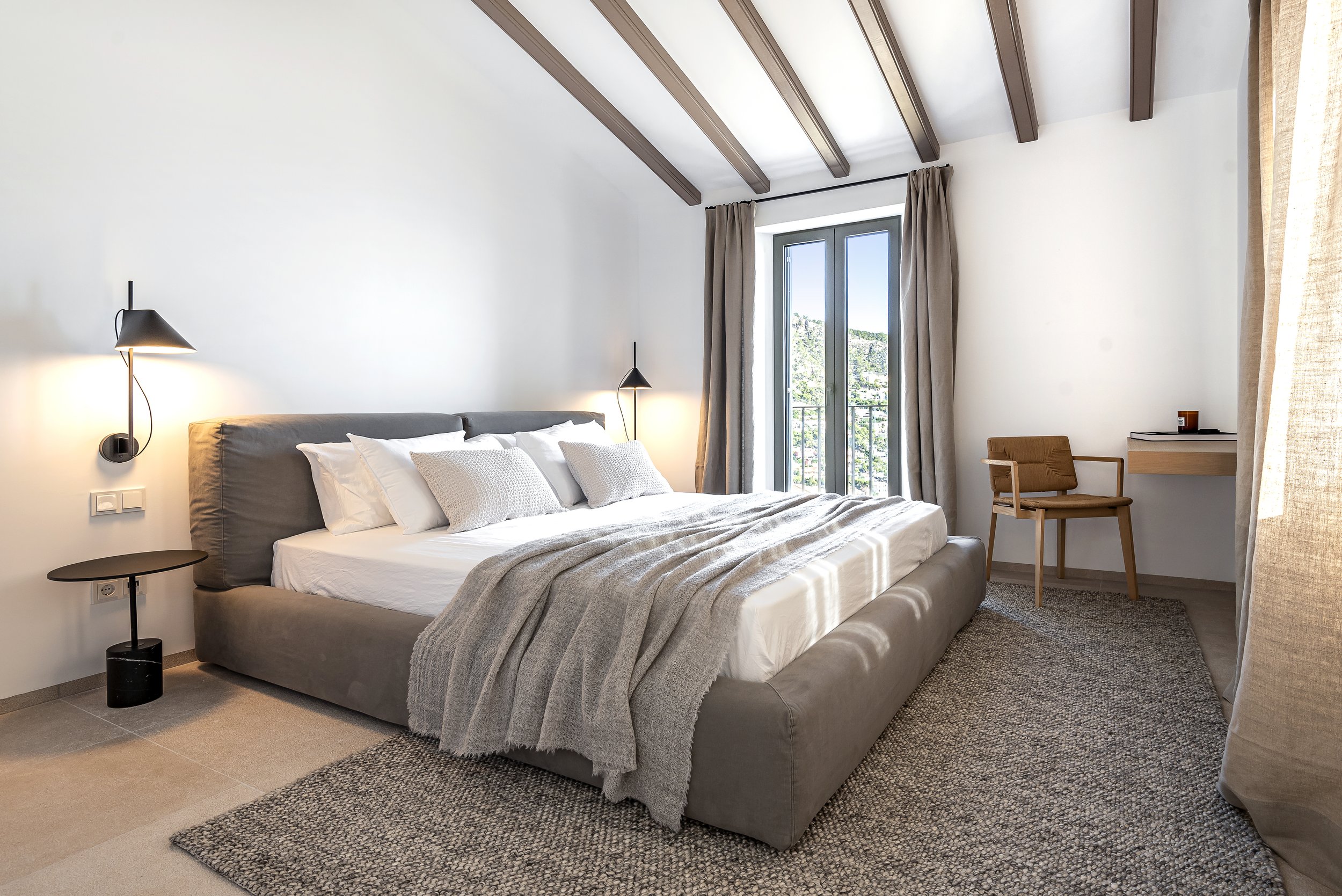 Habitación moderna con cama grande, paredes blancas, ventanas con cortinas beige, lámparas de lectura al lado de la cama, silla de madera en una esquina y alfombra de yute.