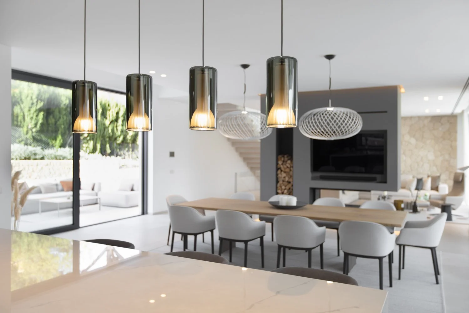 Interior de una sala moderna con lámparas colgantes de diseño elegante, mesas y sillas blancas, una sala de estar con sillones y una vista al exterior a través de grandes ventanas.