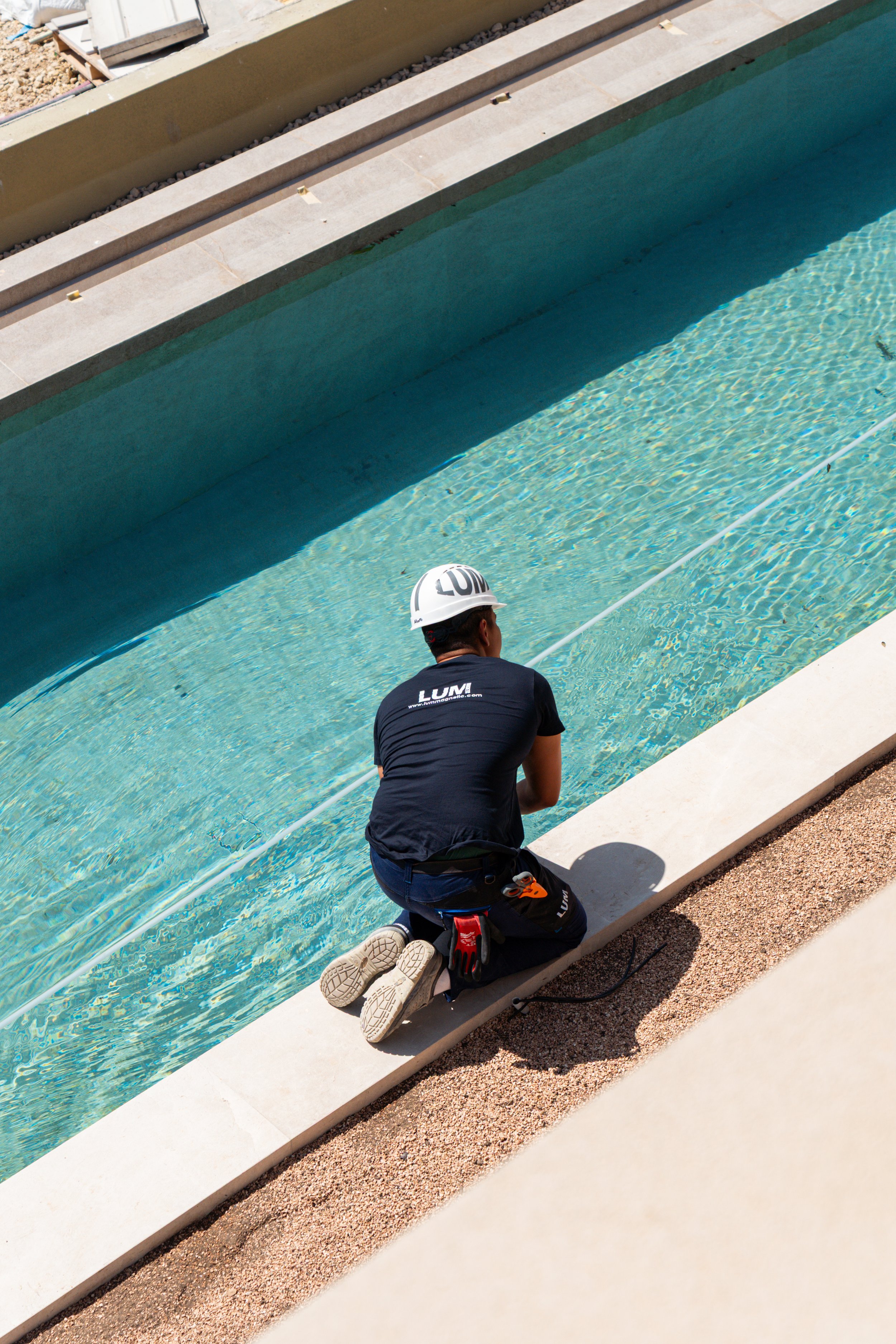 Trabajador con casco y camiseta negra con logo LUM, kneeling junto a la piscina, con herramientas y equipo de construcción a su alrededor.