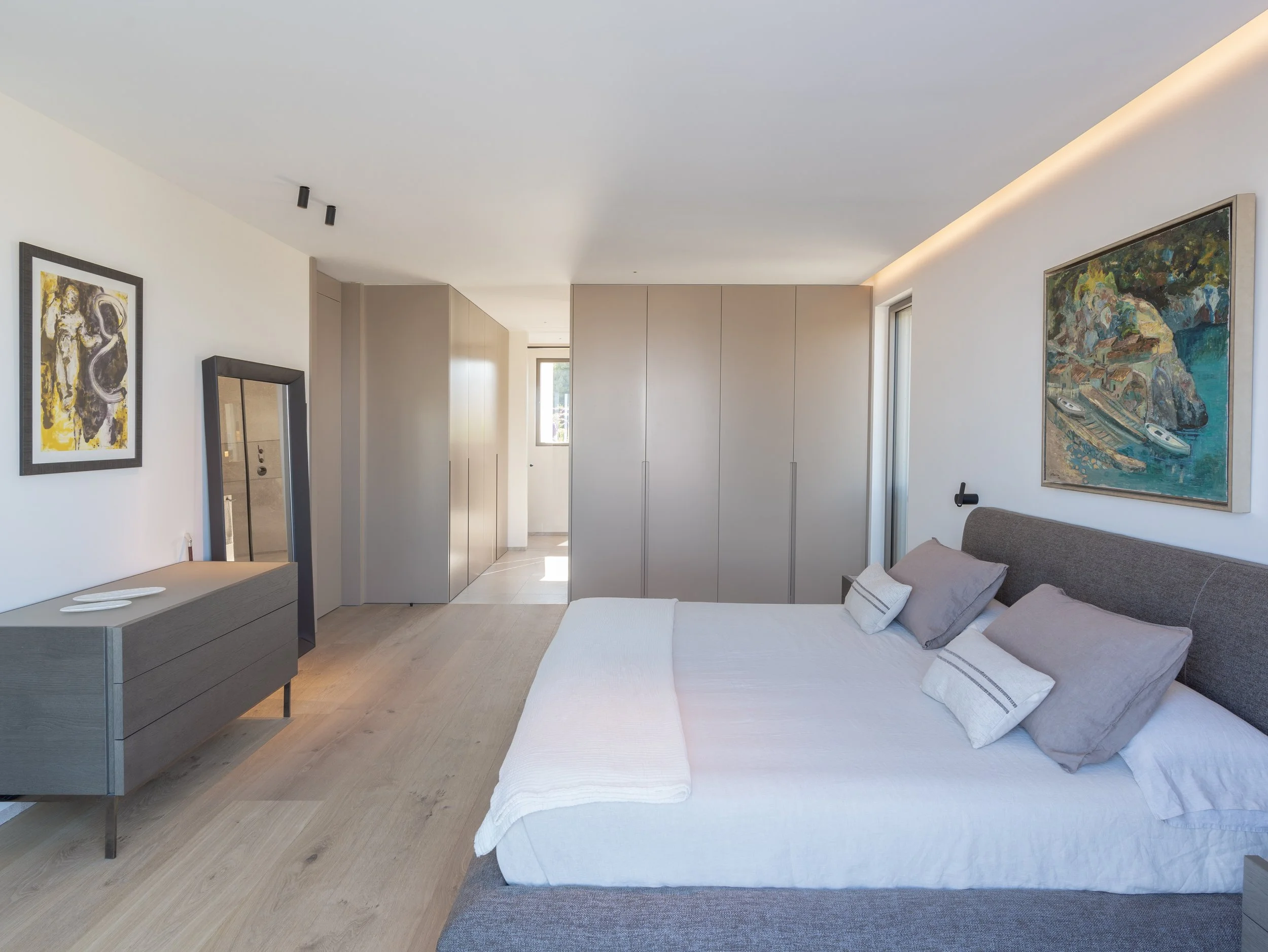 Dormitorio moderno con paredes blancas, arte en las paredes, una cama con sábanas blancas y almohadas, y una ventana que deja entrar luz natural.