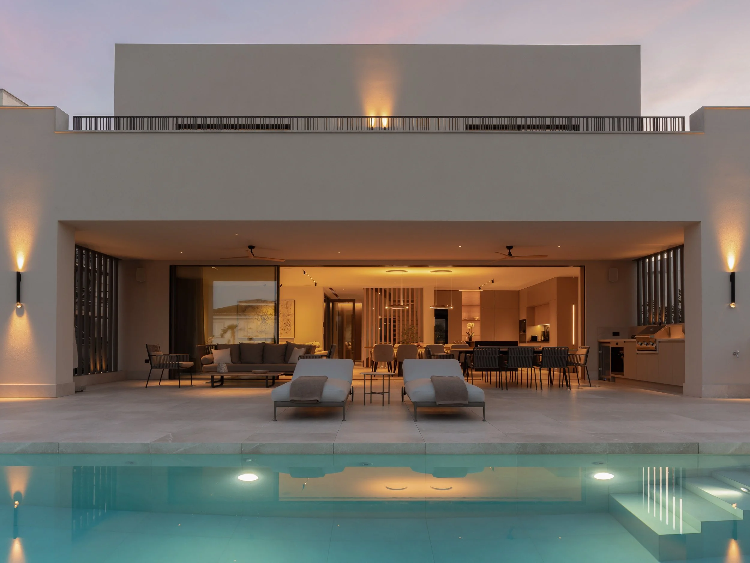 Vista frontal de una terraza de diseño moderno con muebles contemporáneos, techo con ventiladores y espacio interior visible desde afuera, con piscina reflectante en primer plano.