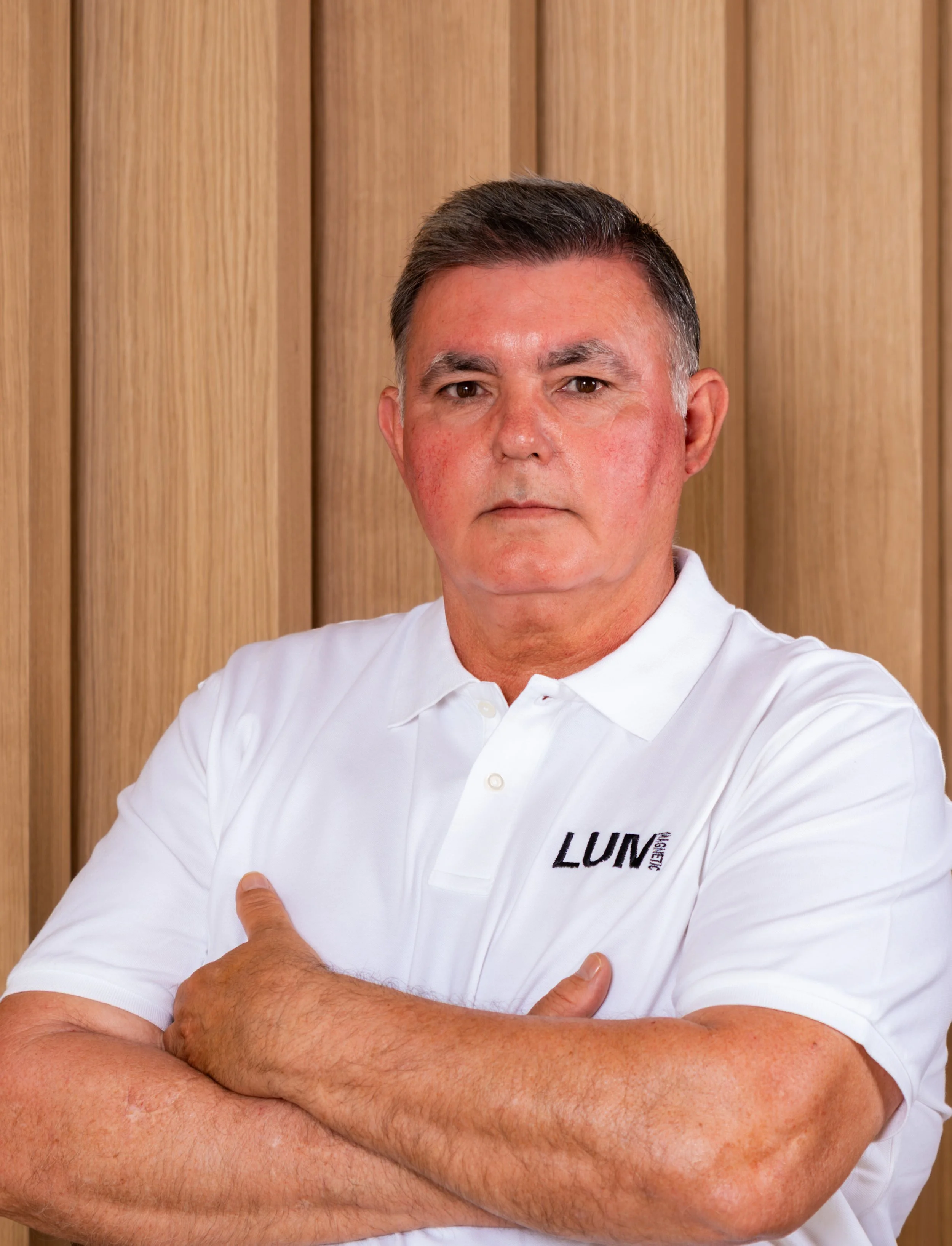 Hombre con camisa blanca que tiene la palabra LUM en el pecho, con los brazos cruzados frente a un fondo de madera.