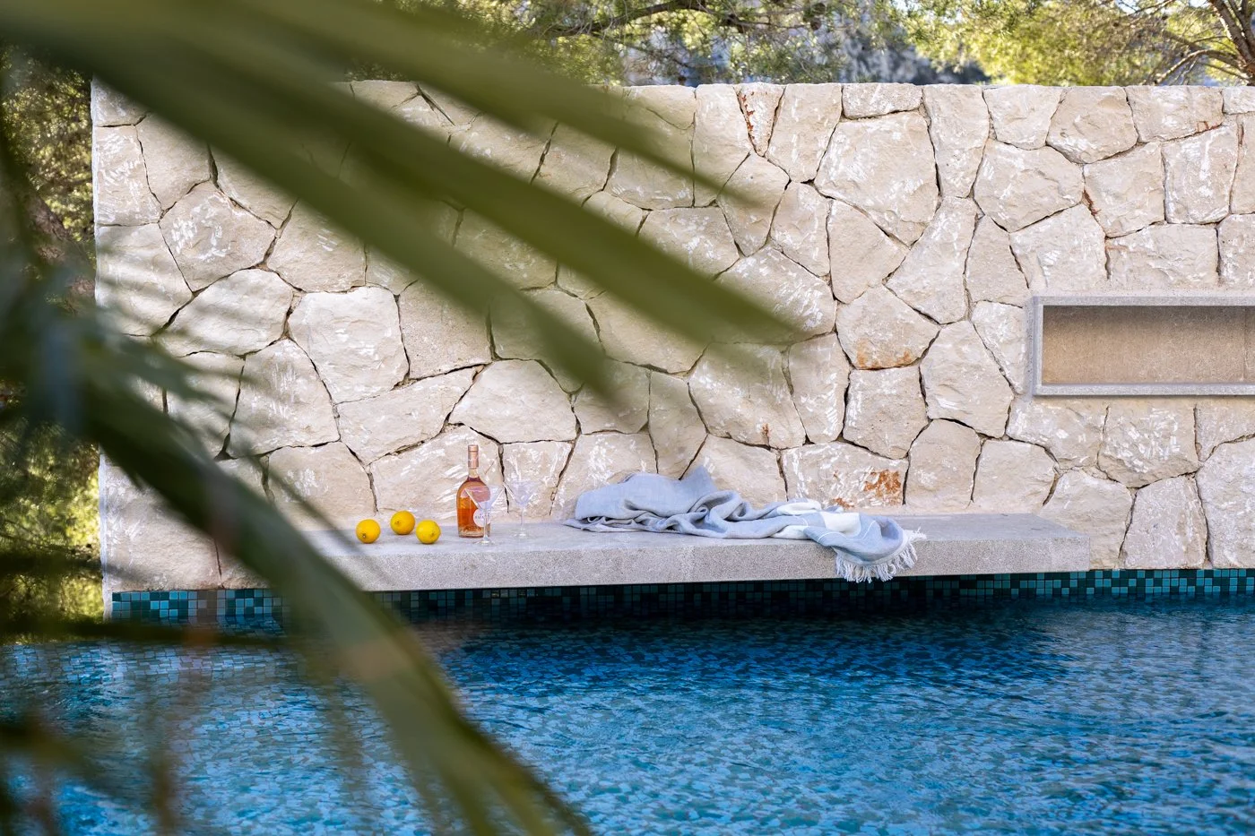 Pared de piedra junto a una piscina con objetos de verano, incluyendo una botella de rosé, tres limones, una toalla gris y una copa, con vegetación en primer plano.