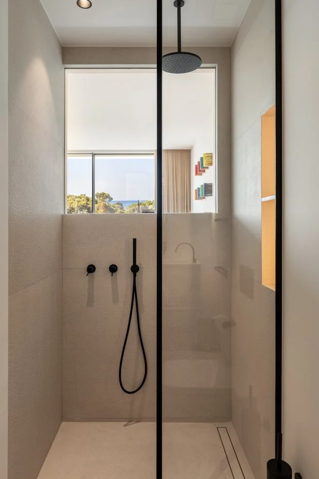 Ducha moderna con paredes de piedra, ducha de lluvia, vista a través de un ventanal grande con vista al exterior, y un espejo que refleja una pared decorada con cuadros coloridos.