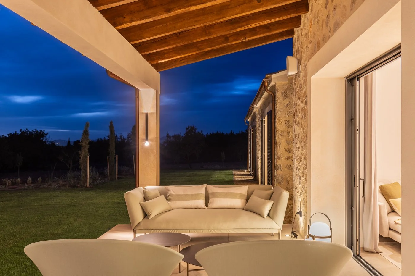 Sala de estar exterior con sofá beige, sillones blancos, mesa y lámpara, vista al campo durante la noche.