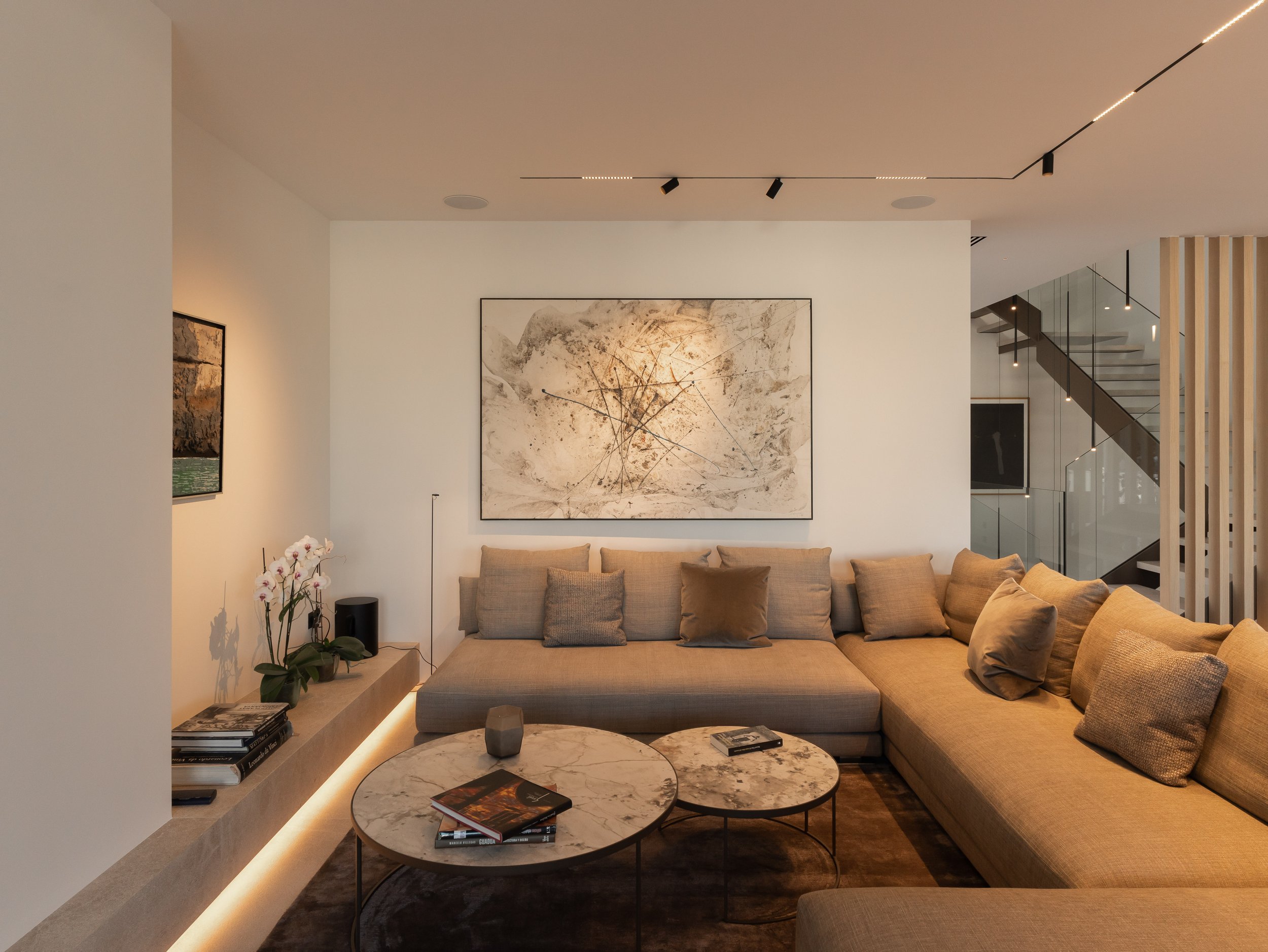 Sala de estar moderna con un sofá beige, mesa de centro de mármol con libros y revistas, pared blanca con arte abstracto, palmera ornamental y escaleras de vidrio y madera.