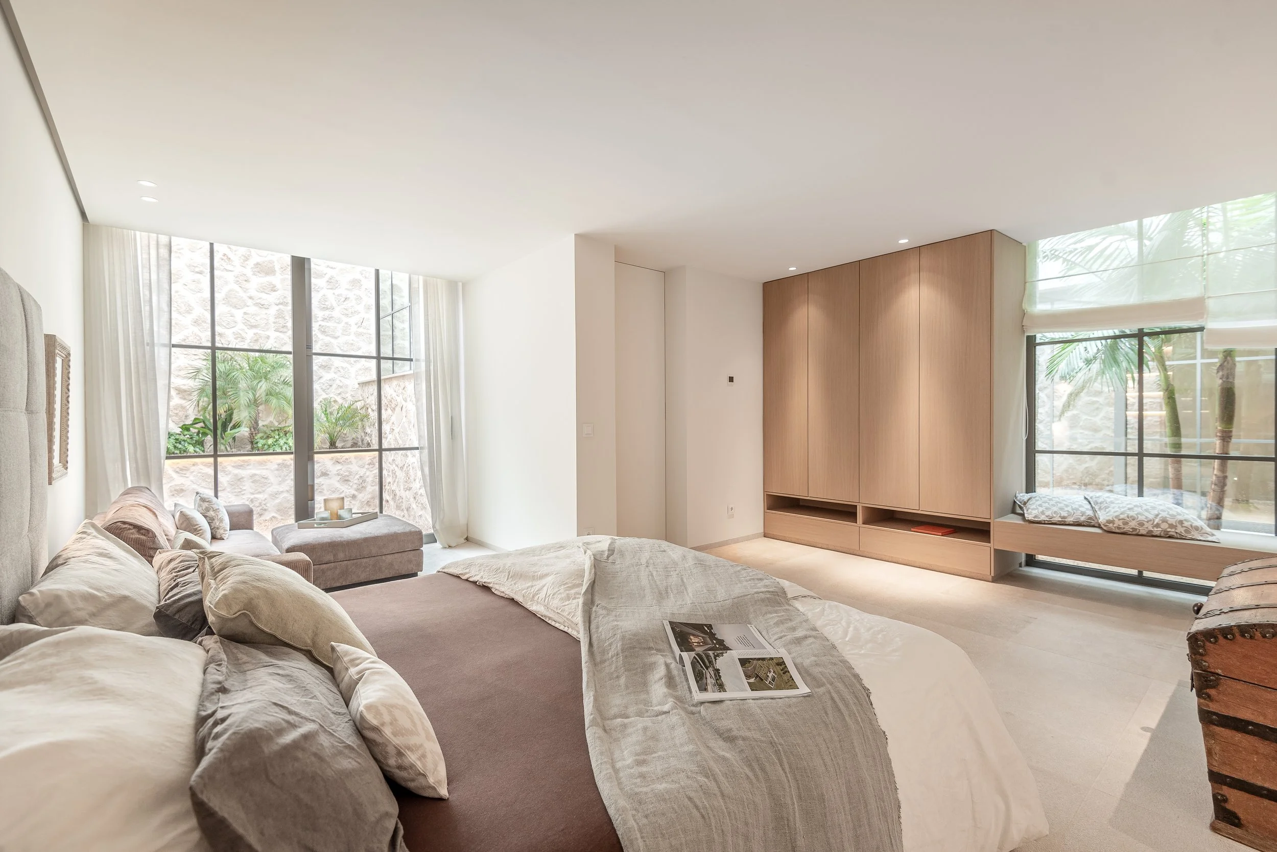 Sala de estar con grandes ventanas, sillones beige, una cama con almohadas, y una mesa con revistas, decorada con muebles de madera y cortinas blancas.