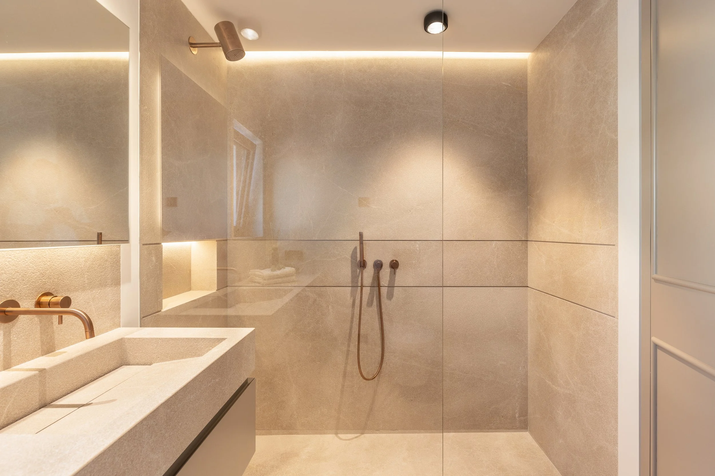 Una ducha moderna con paredes y suelo de azulejos beige, ducha con cabezal de lluvia y accesorios de cobre, espejo y lavabo en primer plano.