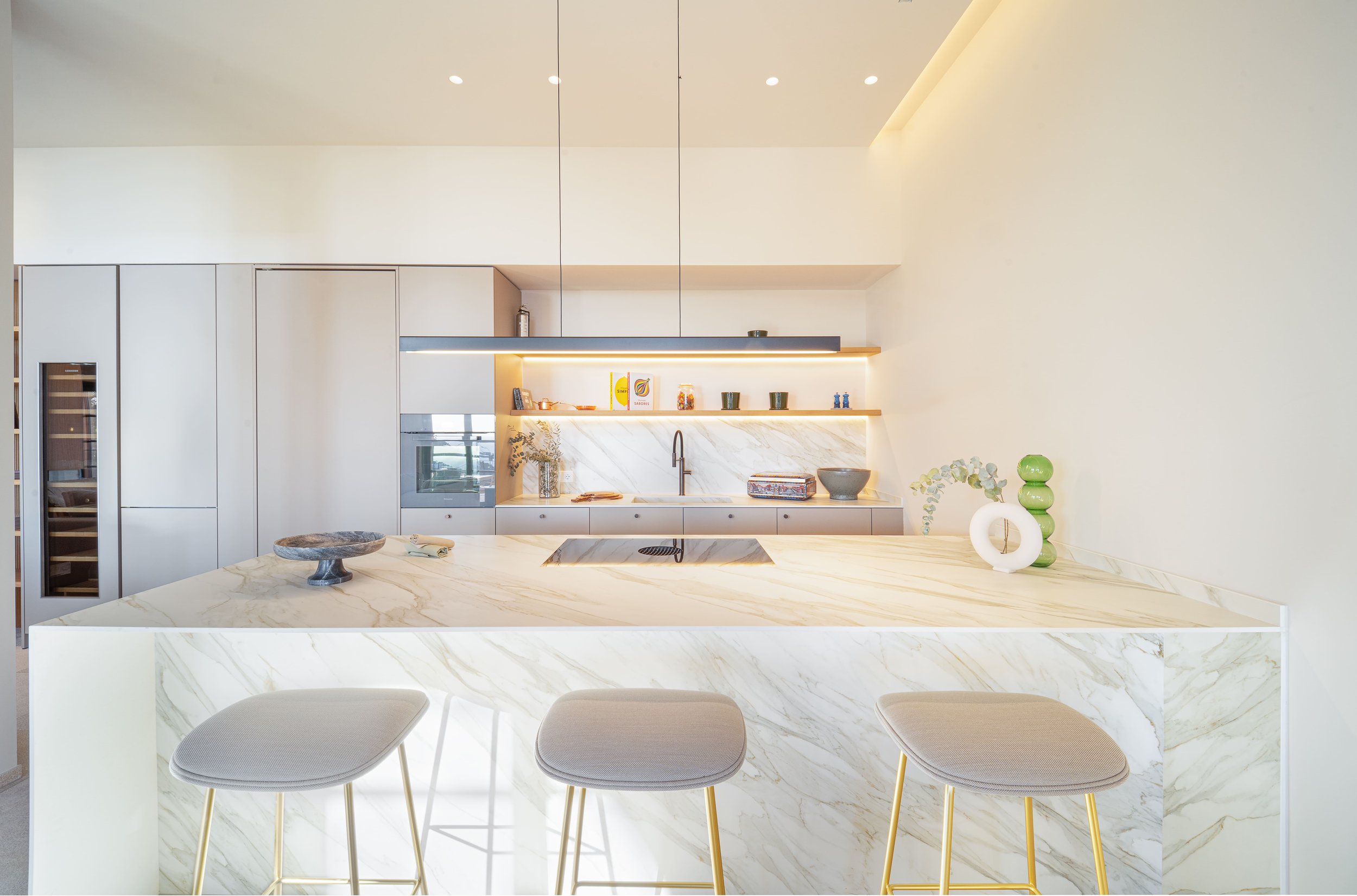 Cocina moderna con encimera de mármol, armarios en tonos claros, estantería con decoraciones y utensilios, y sillas en la zona de desayuno.