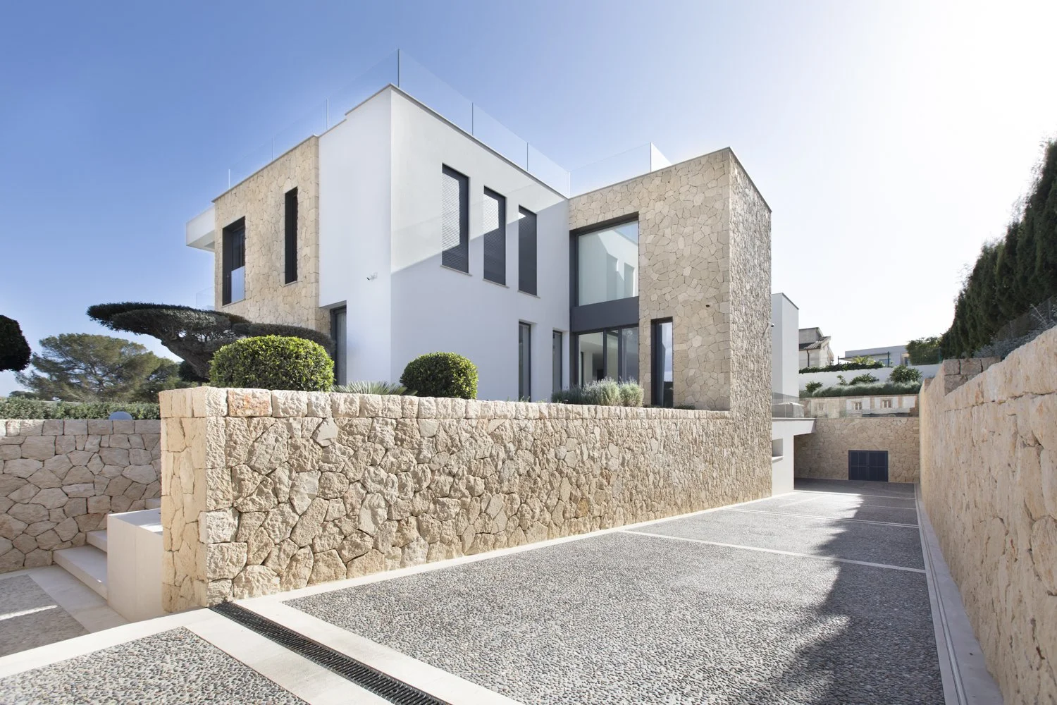 Vista frontal de una casa moderna de varios pisos con paredes de piedra y blancas, ventanas altas y un área de estacionamiento pavimentada con muro de piedra en el entorno.