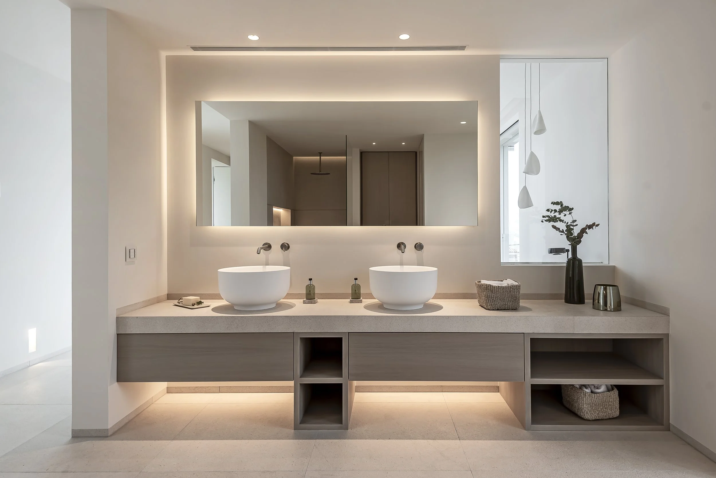 Baño moderno con dos tinas blancas y un gran espejo con iluminación LED en una pared. Hay una ventana y decoraciones minimalistas, incluyendo plantas y una cesta con toallas.