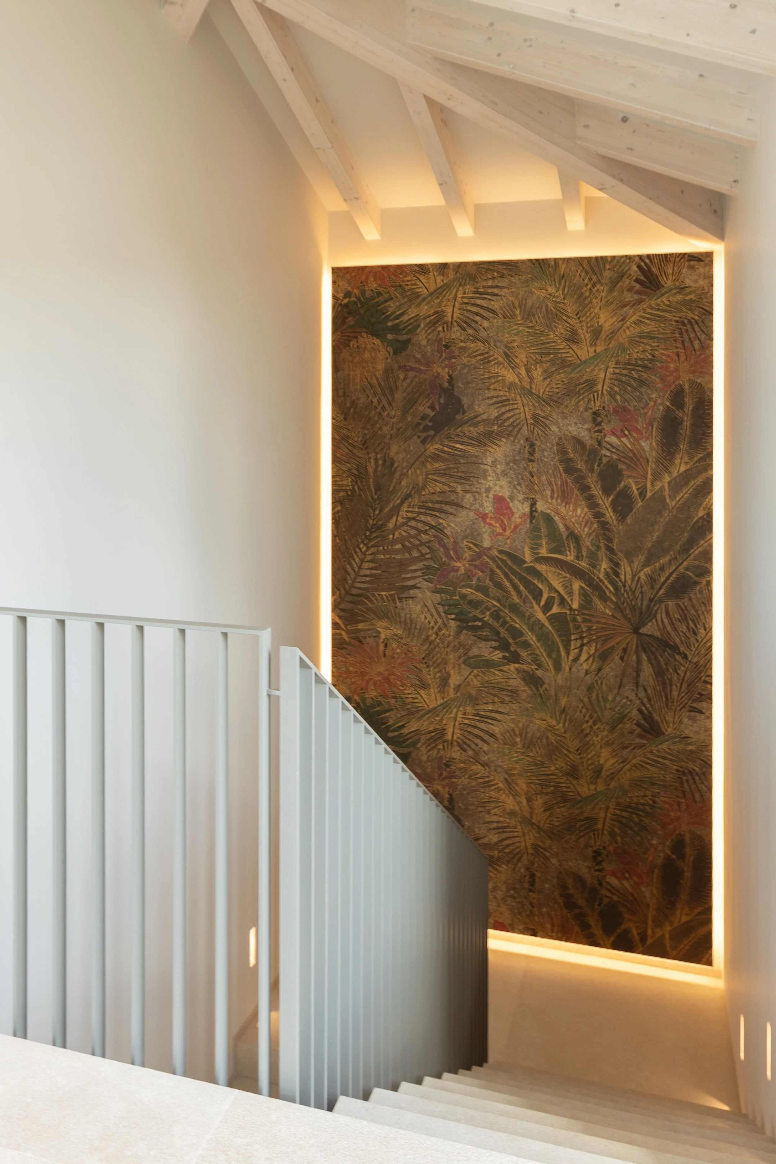 Escalera moderna con barandal blanco que baja hacia un pasillo iluminado, con una pared decorativa con un mural de plantas tropicales y iluminación en los bordes.