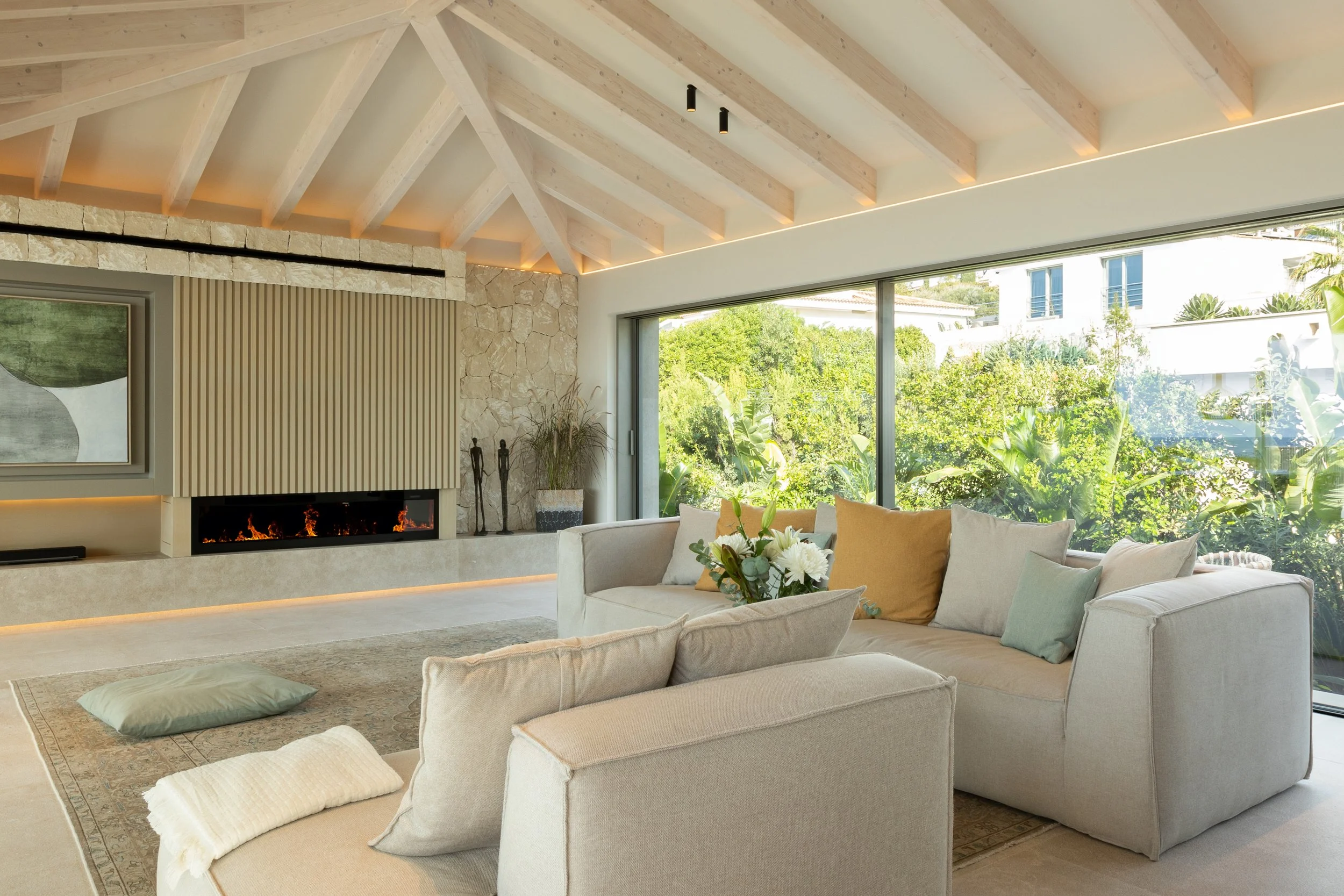 Salón con techo de vigas de madera y vista a un jardín exterior con árboles y plantas.