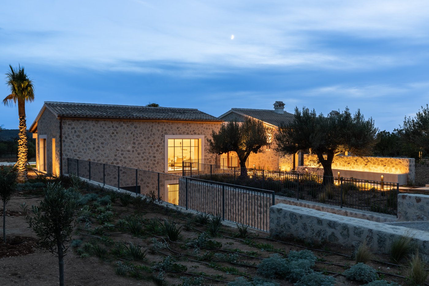 Casa moderna de piedra con árboles y jardines, iluminada en la noche.