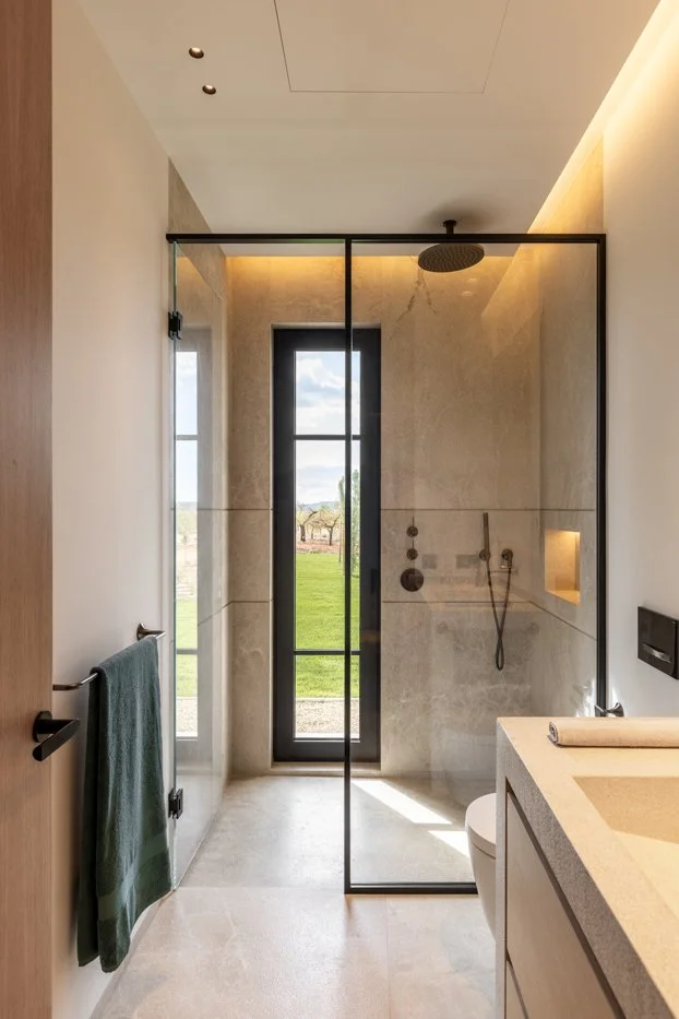 Baño moderno con ducha de cristal, ventana grande con vista a un campo verde, lavabo y lavabo pequeño, toalla verde colgada, iluminación cálida en el techo.