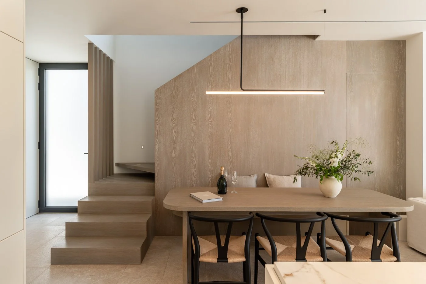Sala de comedor moderna con mesa de madera, sillas negras y una lámpara lineal suspendida, decorada con una planta en un jarrón y una botella de vino con copas