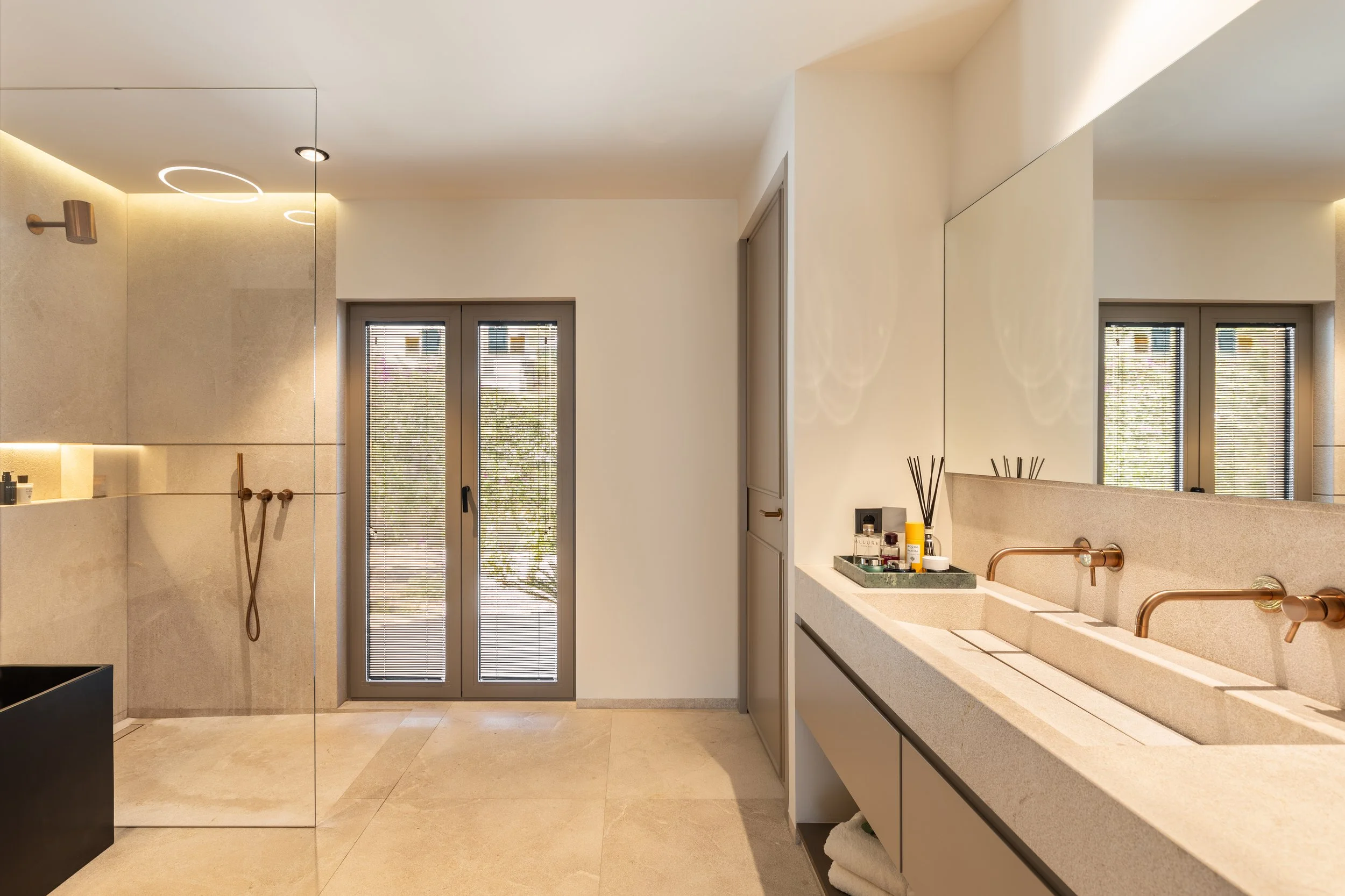 Baño moderno con lavabos de piedra, grandes ventanas con persianas, ducha con ducha de mano, decoraciones en la encimera, paredes de color claro y pisos de piedra beige.
