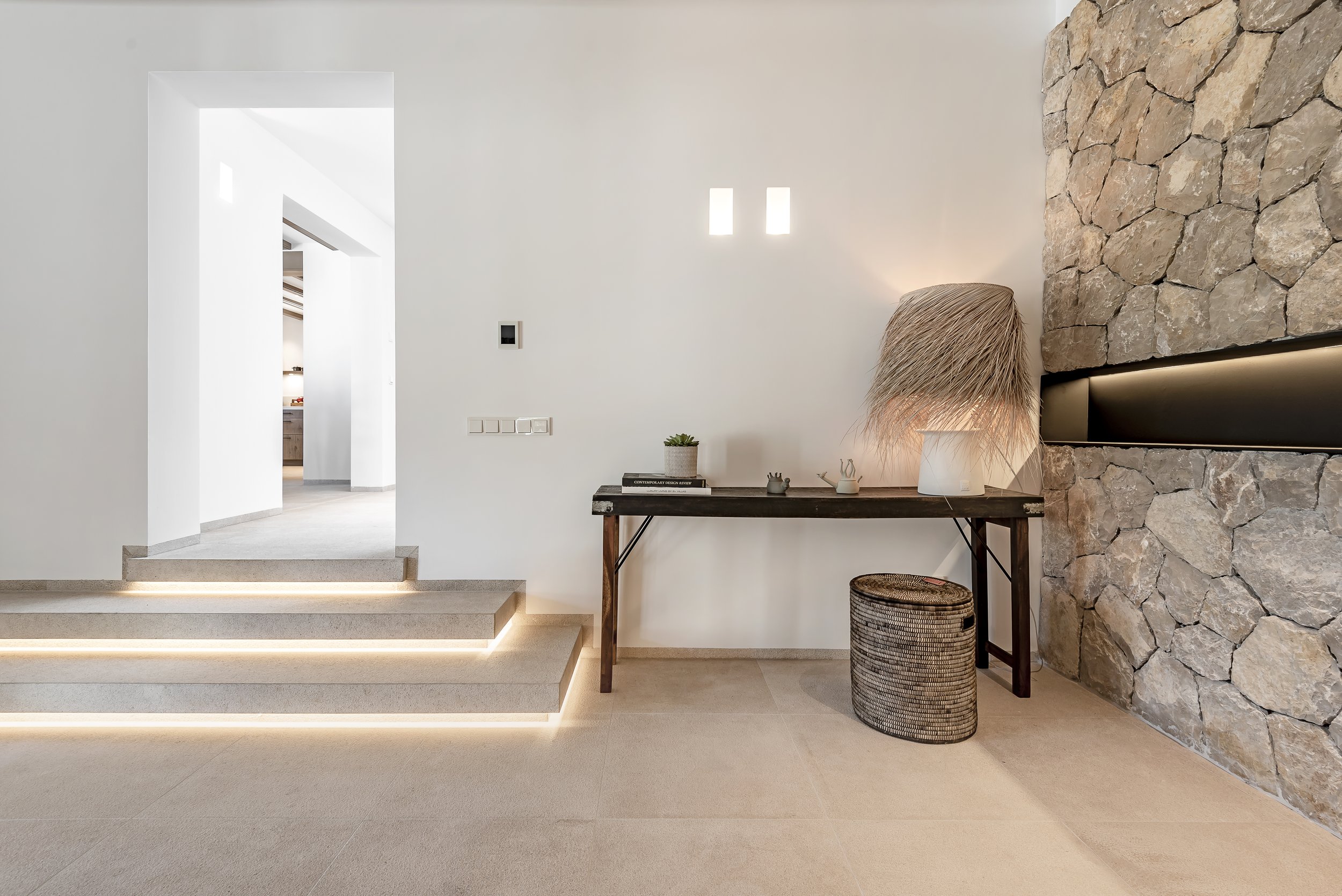 Interiores modernos con paredes blancas y decoraciones en tonos neutros, con una pared de piedra y una mesa con objetos decorativos y plantas.